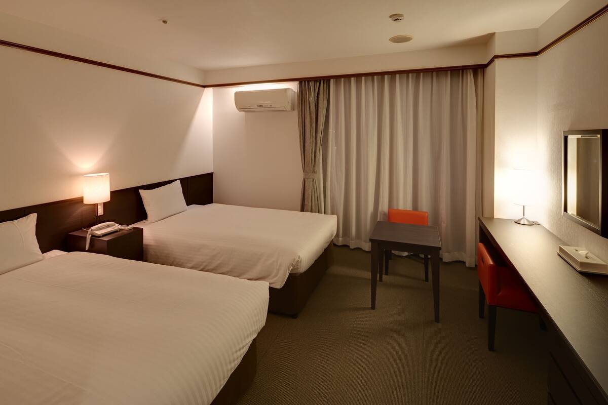 Ein Hotelzimmer mit zwei Betten, einem Schreibtisch, einem Stuhl, einem Fernseher und einer Klimaanlage.