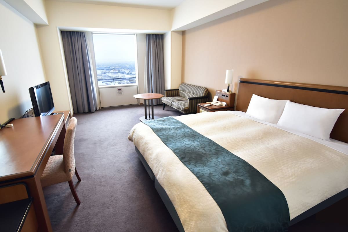 A hotel room with a bed, a desk, a chair, a small table, and a view of the city through the window.