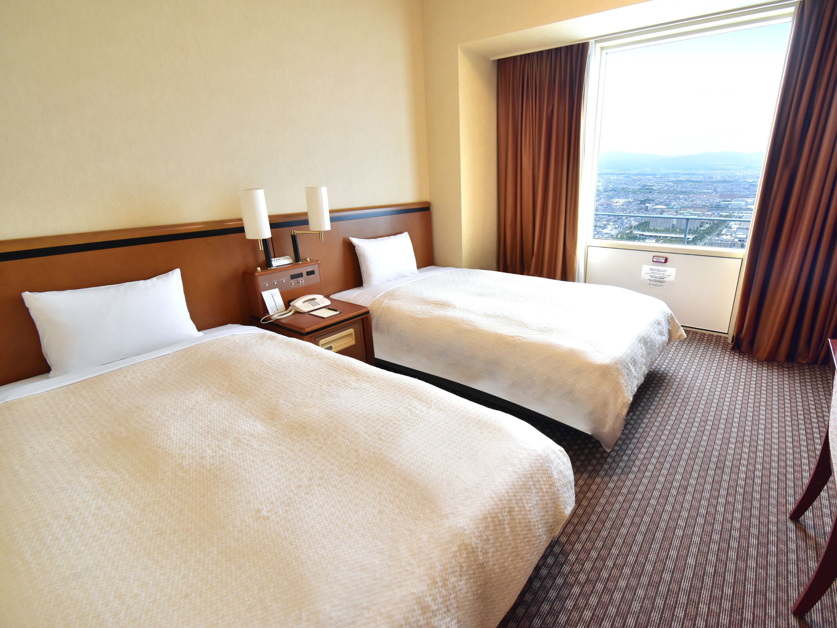 Een hotelkamer met twee bedden, een nachtkastje met telefoon en een raam met uitzicht op de stad.