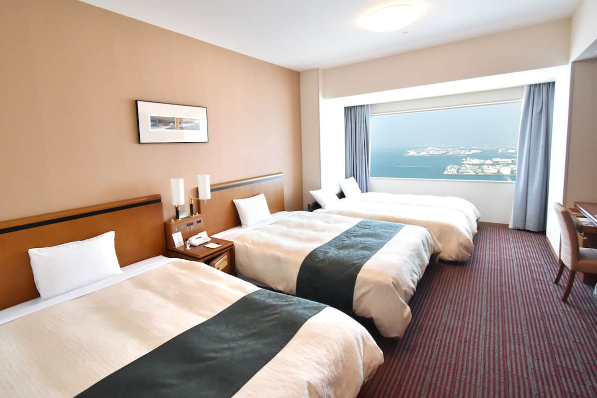 A hotel room with two beds, a desk, a chair, a nightstand, a lamp, a phone, a picture on the wall, and a large window with a view of the city.