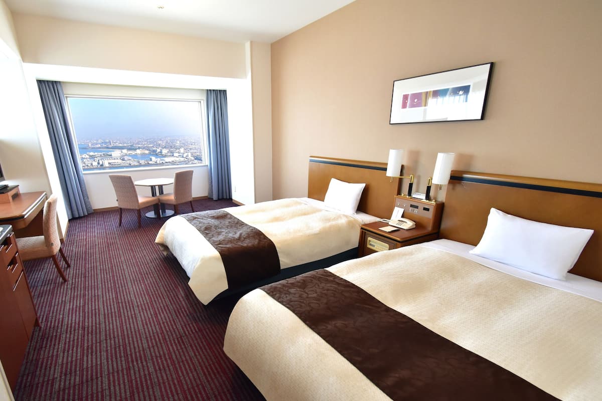 A hotel room with two beds, a desk, a chair, a small table, a nightstand with a phone, and a window with a city view.
