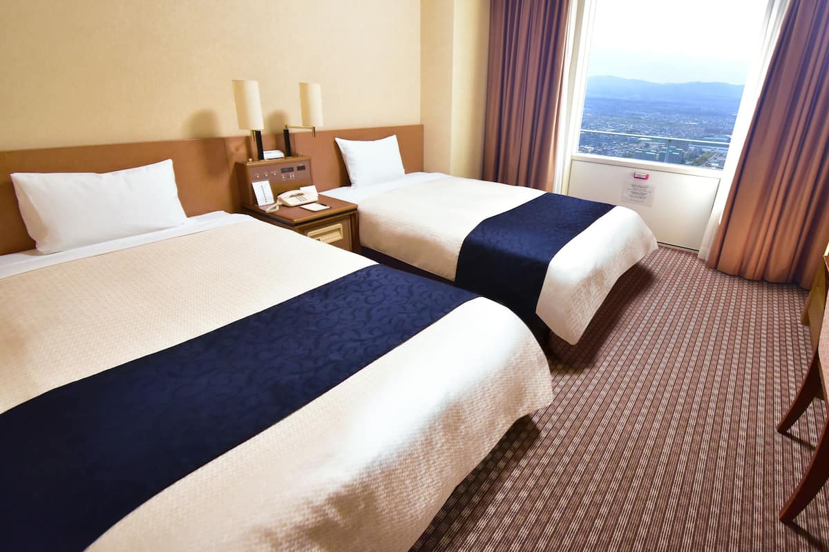 A hotel room with two beds, a nightstand with a phone, and a window with a view of the city.