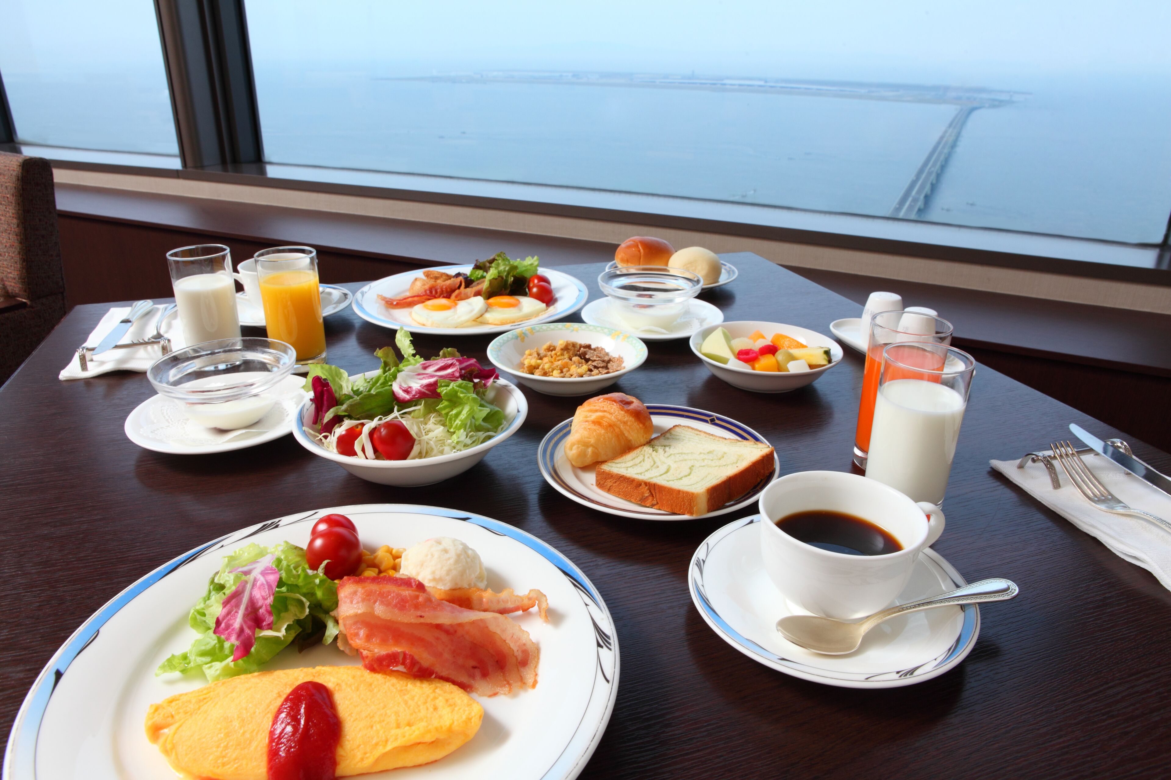 Una mesa de desayuno con diferentes platos, como huevos, tocino, tostadas y fruta, con vista al agua.