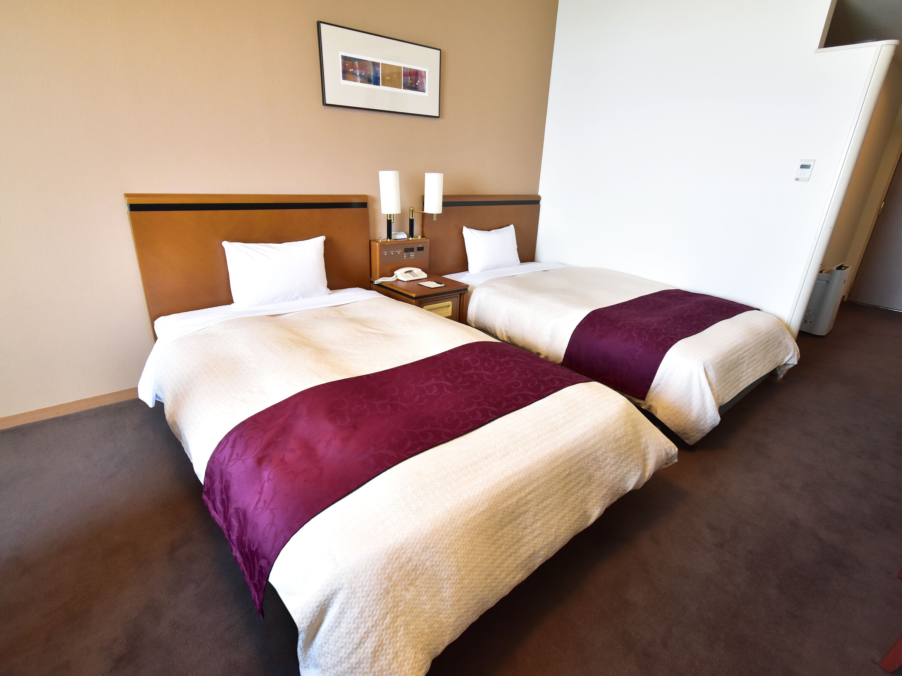 A hotel room with two beds, a nightstand with a phone, a wall-mounted picture, and a white door.
