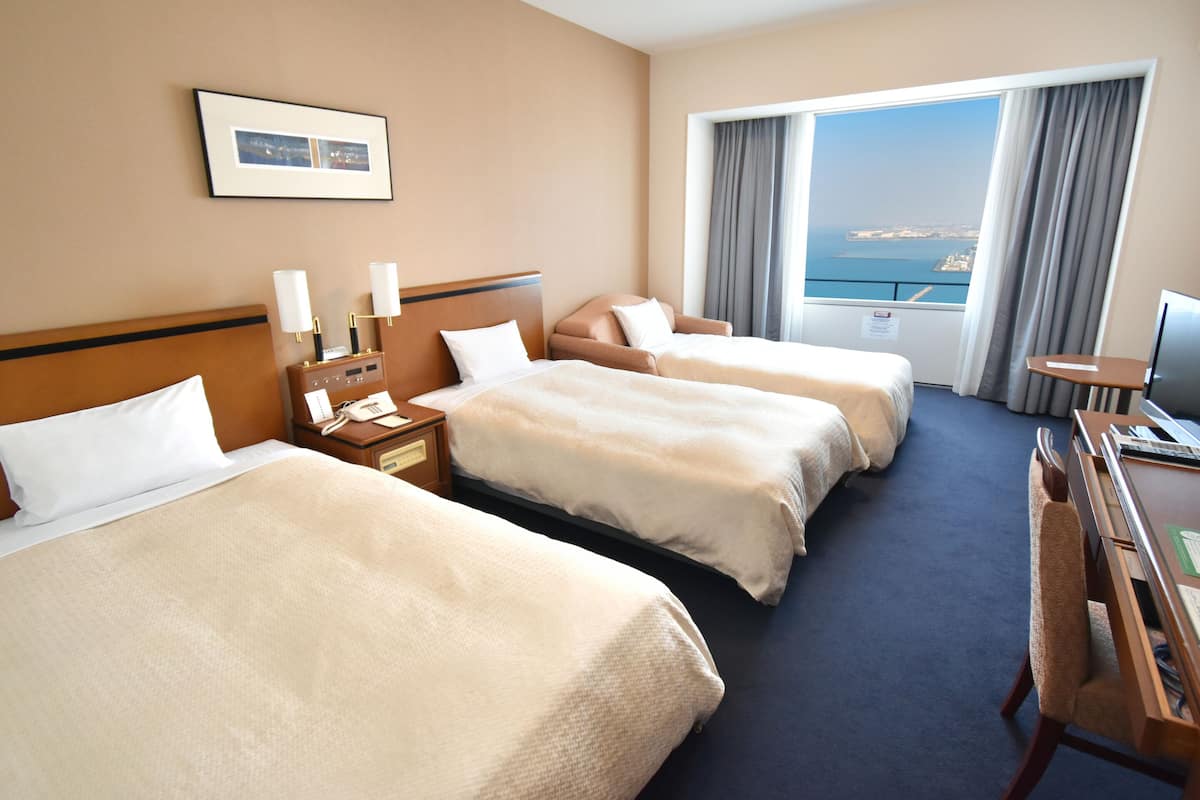 A hotel room with three beds, a desk, a chair, a TV, and a large window with a view of the sea.