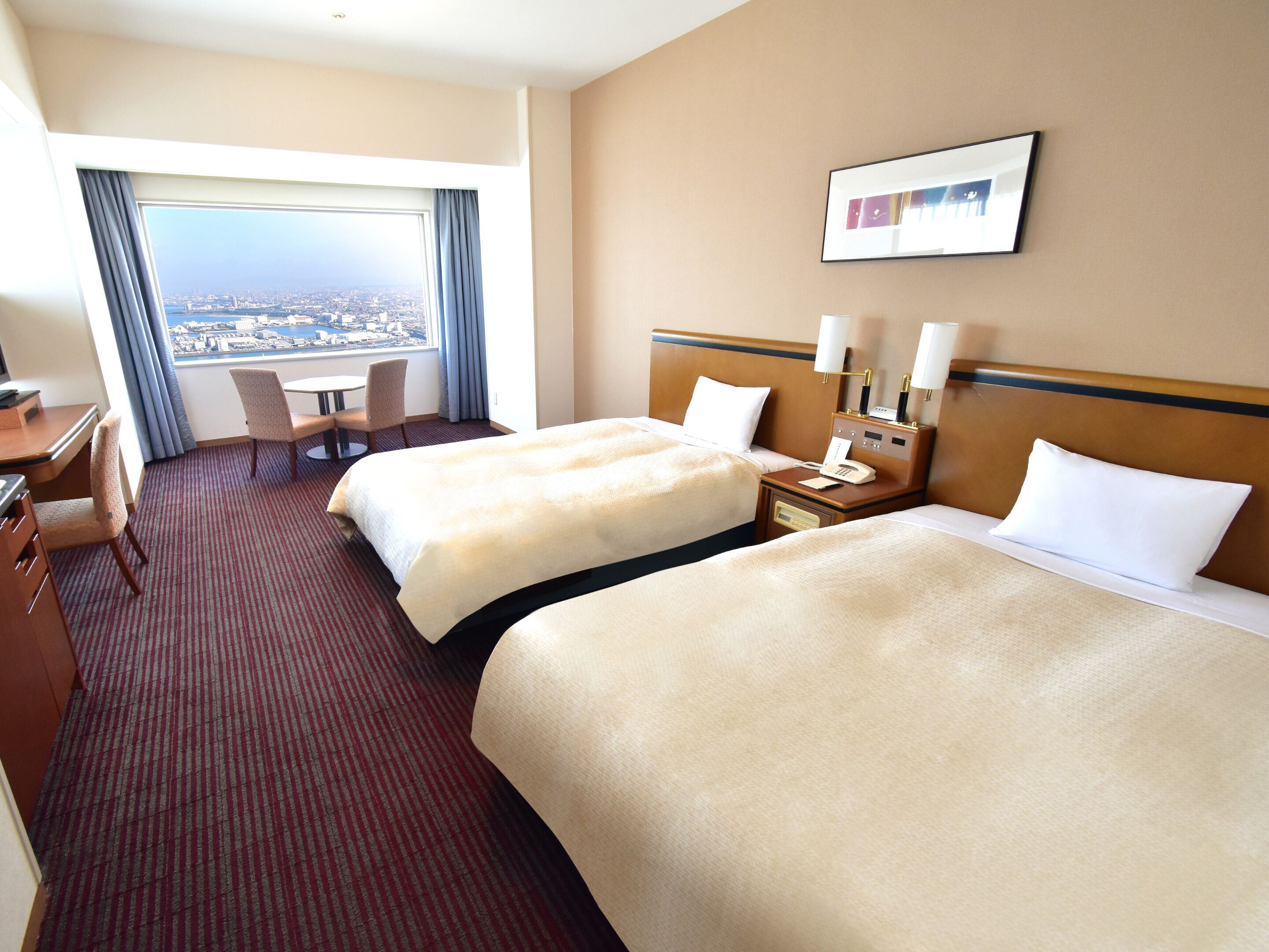 A hotel room with two beds, a desk, a chair, a nightstand with a phone, and a window with a city view.