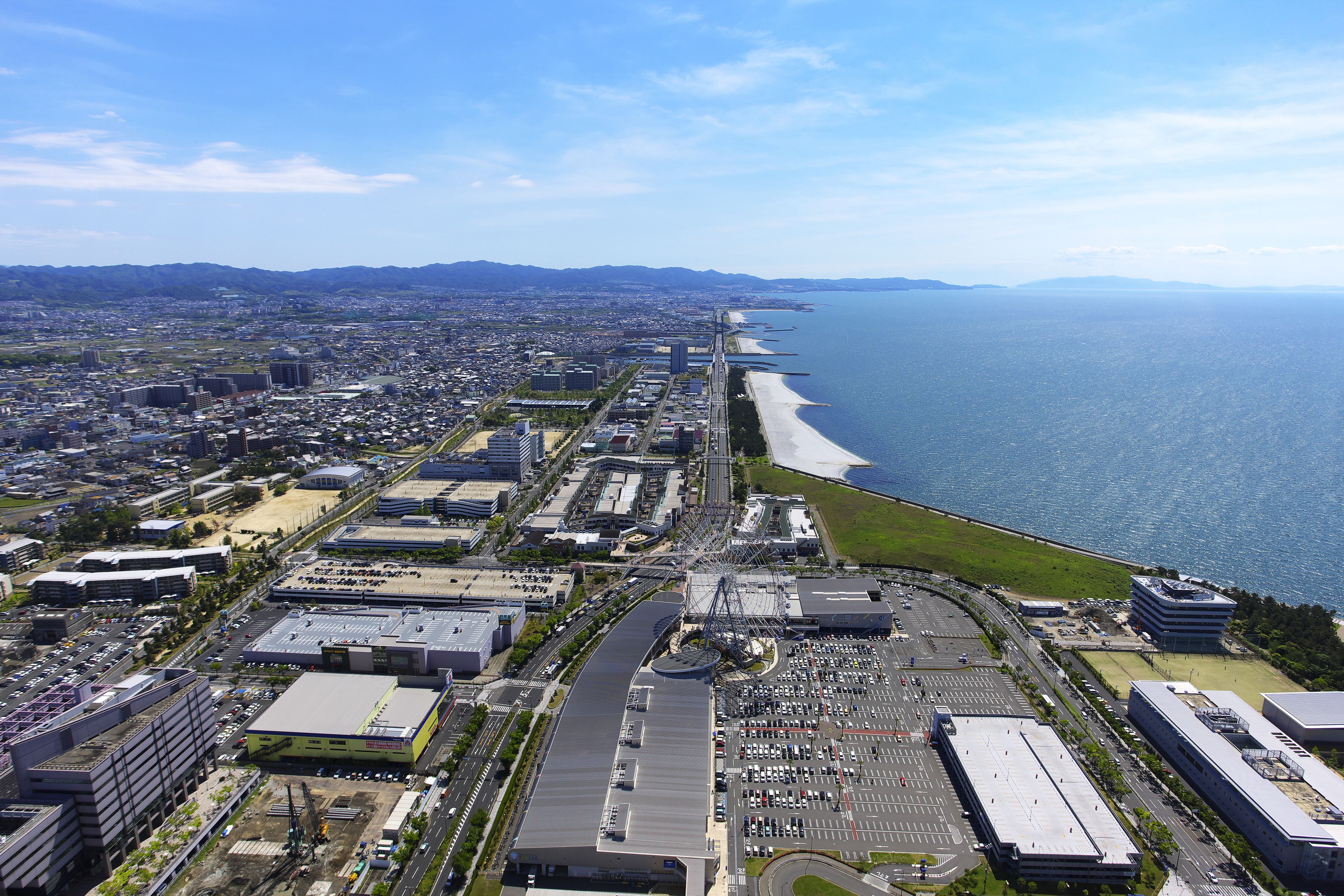 An aerial view of a coastal city with a beach, industrial buildings, and a parking lot.