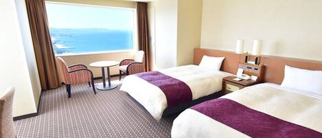 A hotel room with two beds, a small table, two chairs, a nightstand with a lamp, and a view of the sea.