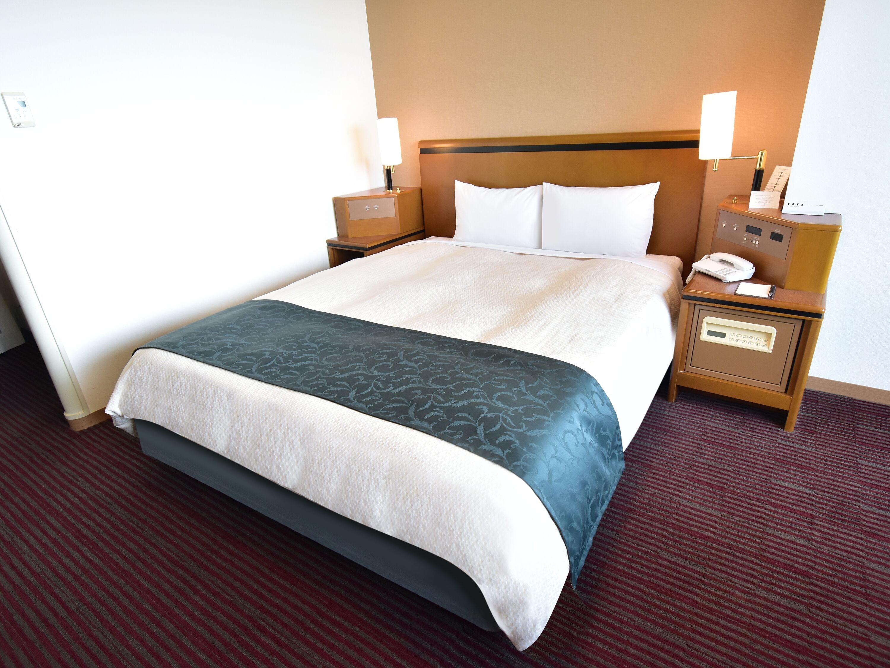 A hotel room with a bed, two bedside tables, a nightstand, a lamp, a phone, and a thermostat.