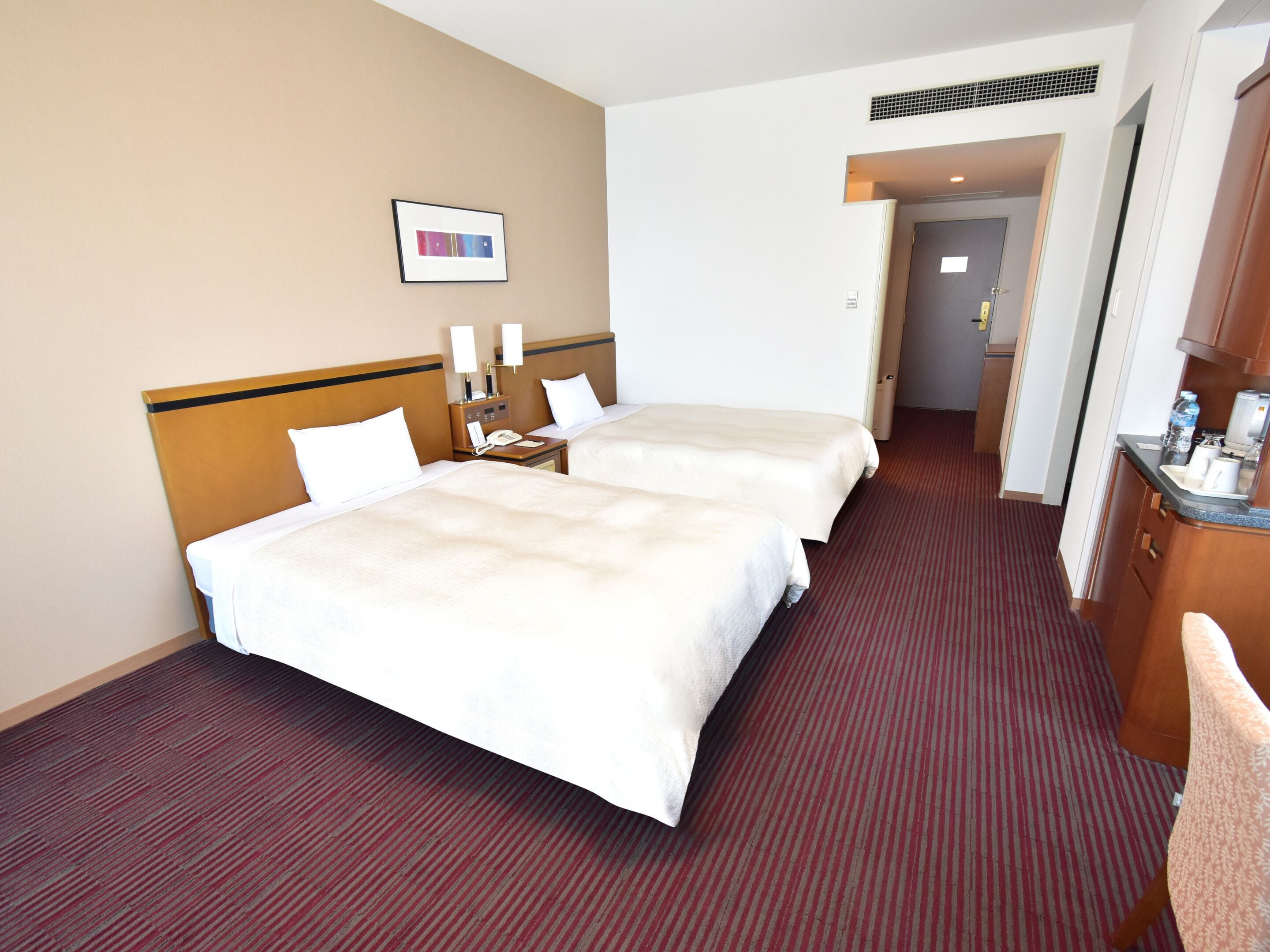 Ein Hotelzimmer mit zwei Betten, einem kleinen Schreibtisch mit Telefon, einem Bild an der Wand und einer Küchenzeile.