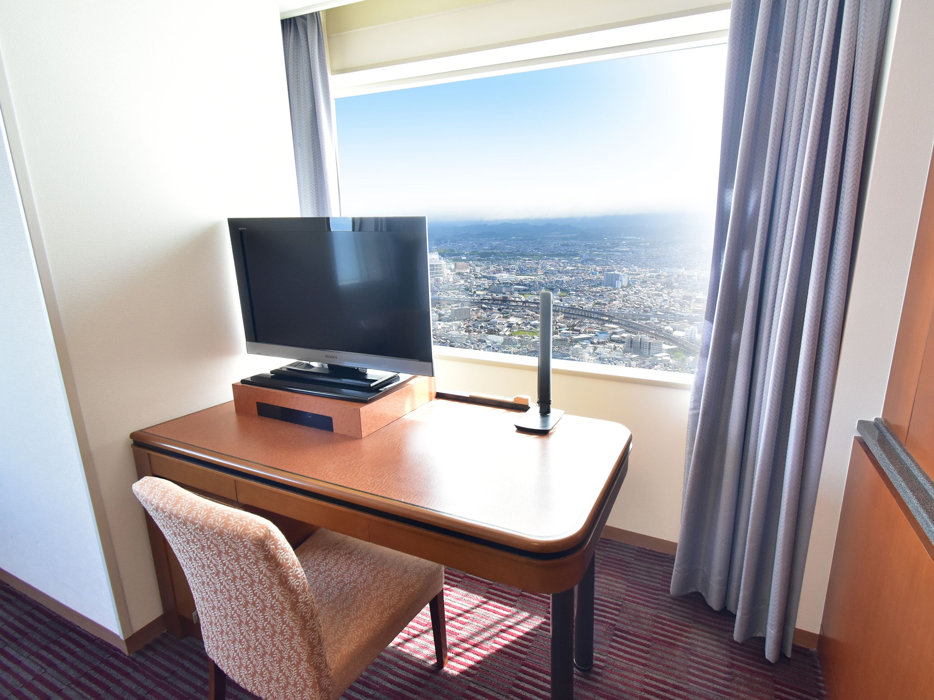 A hotel room with a desk, a flat-screen TV, a chair, and a view of a cityscape through a window.