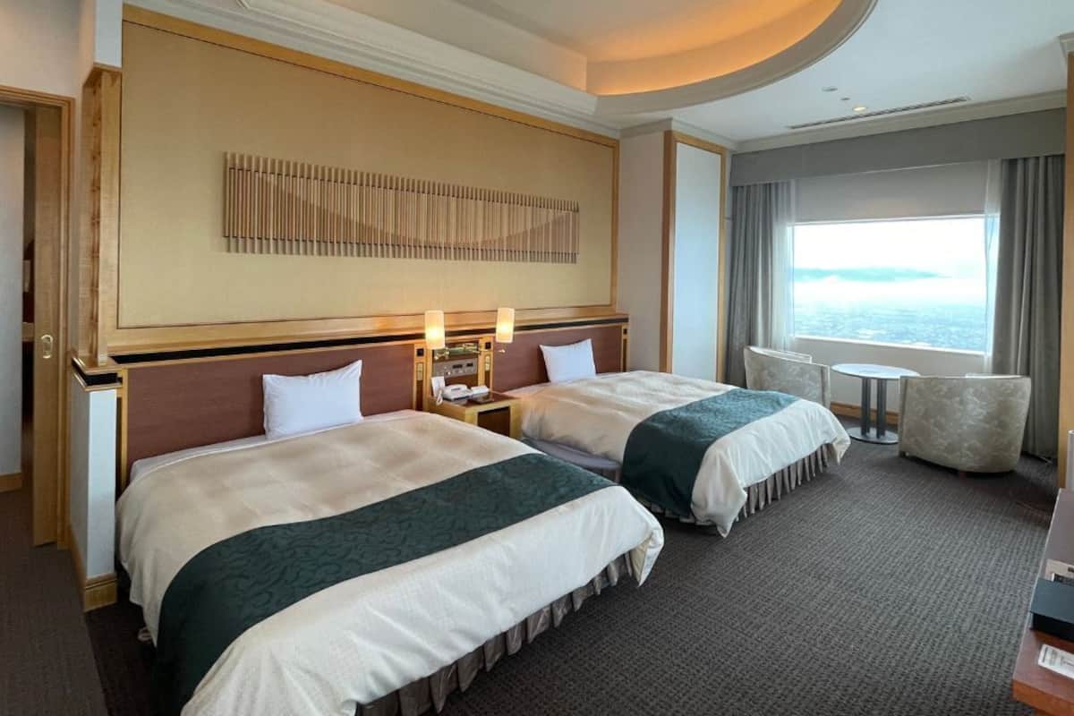 A hotel room with two beds, a large window with a view, a small table, and a chair.