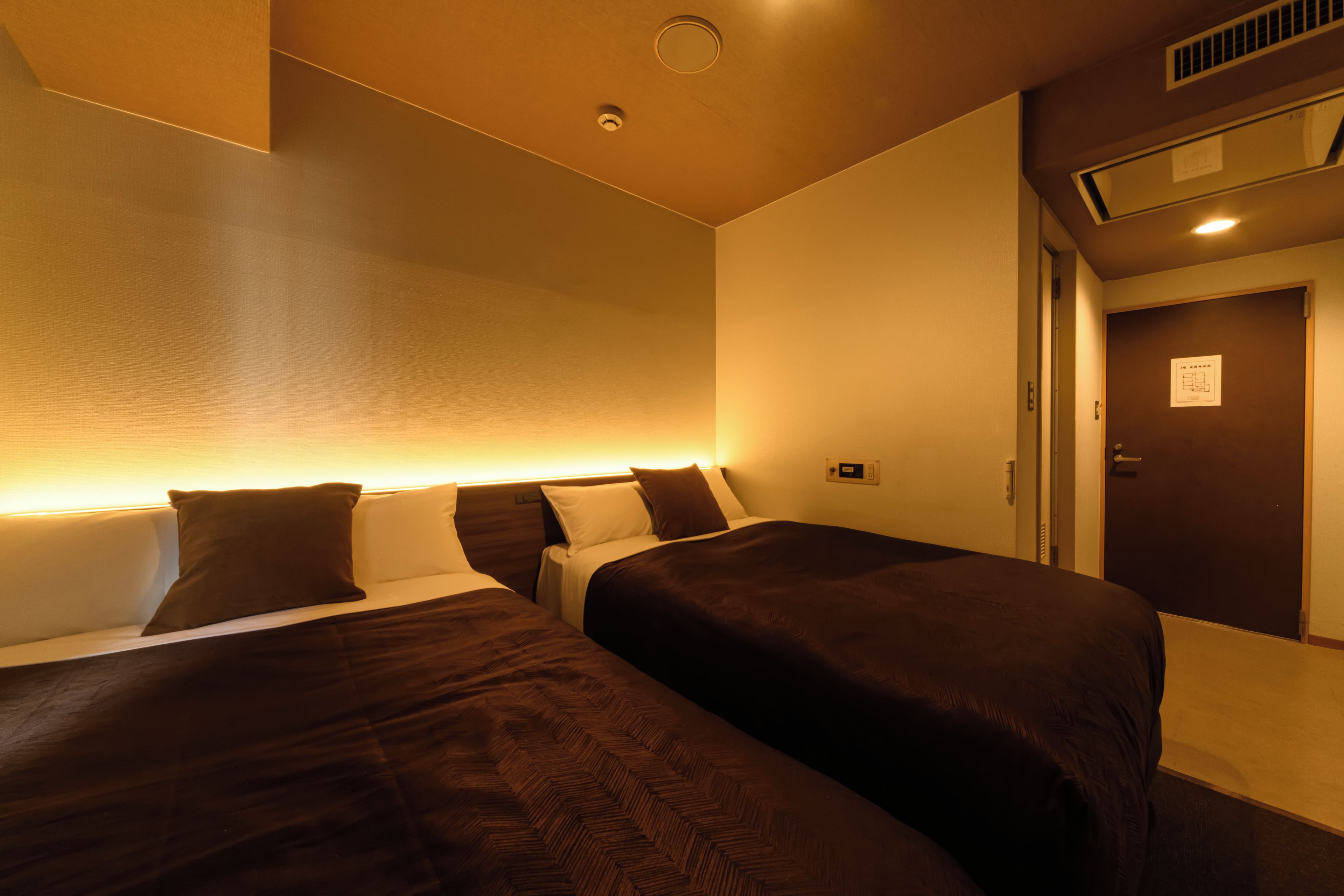 A hotel room with two beds, a wall-mounted heater, a door with a sign, and a ceiling vent.