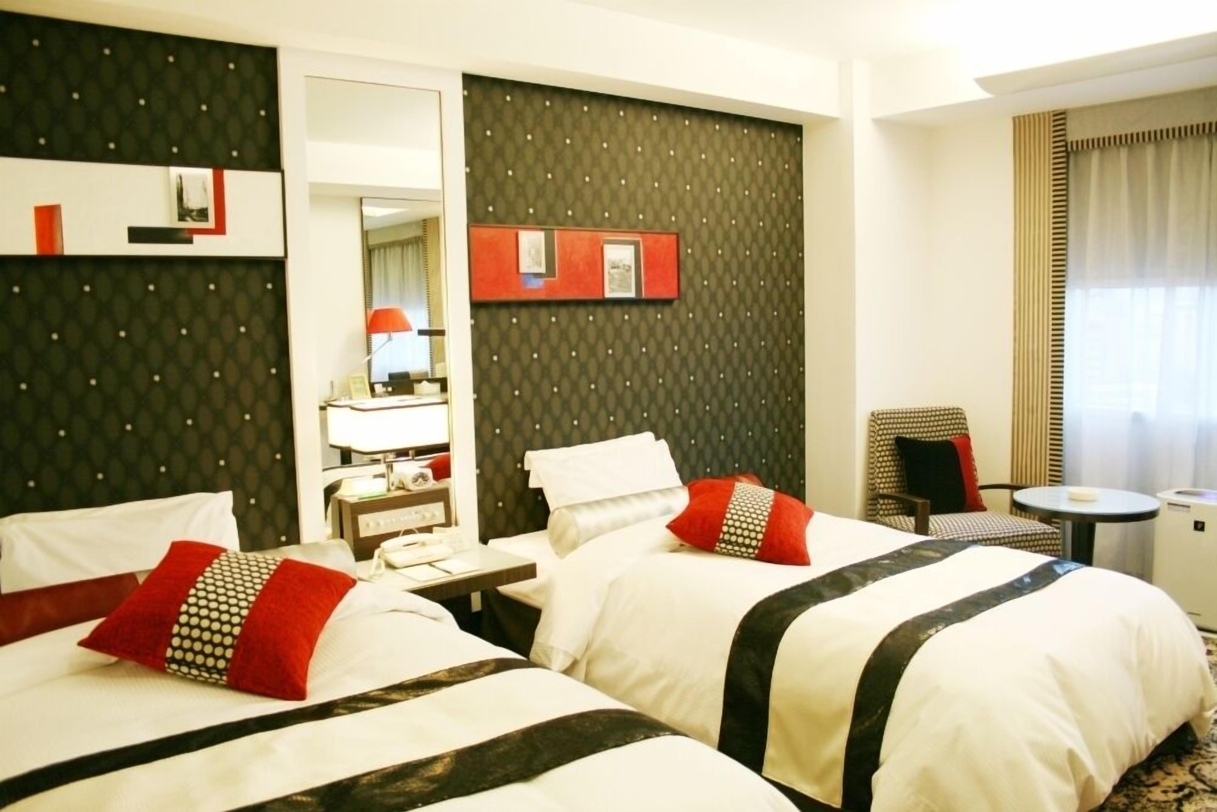 A hotel room with two beds, a small table, a chair, a mirror, and a window with curtains.