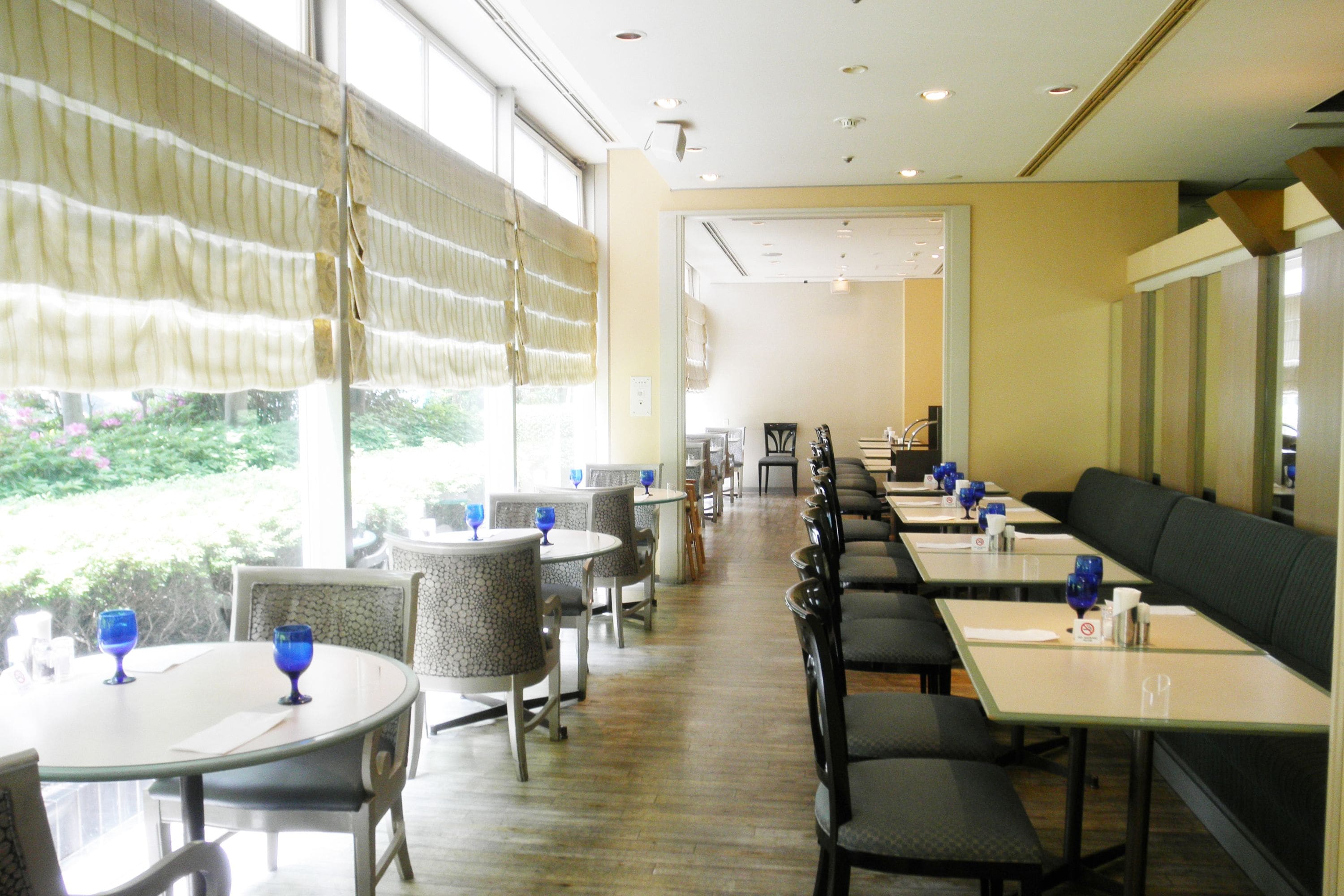 A restaurant with round tables and chairs, a long counter with a black bench, and large windows with curtains.