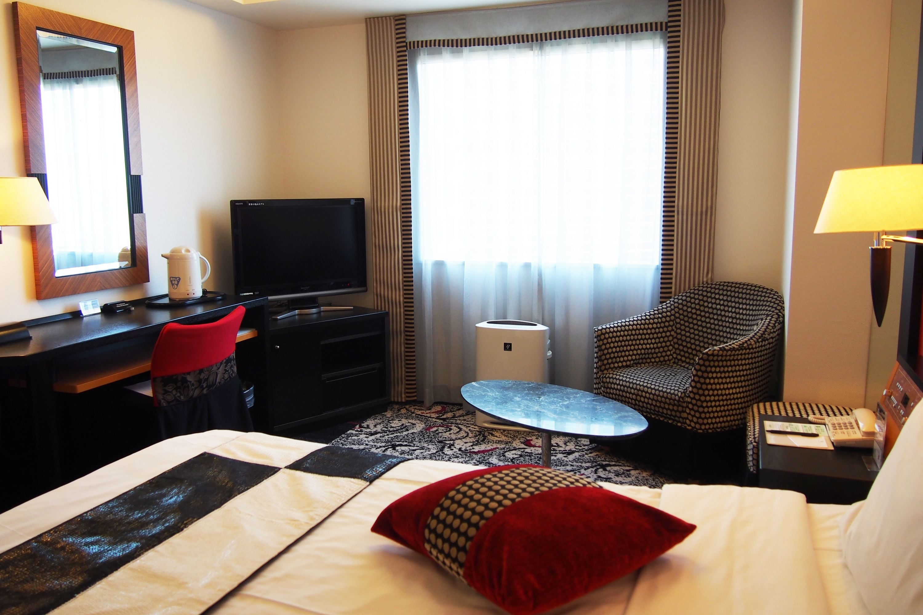 A hotel room with a bed, a desk, a television, a chair, a small table, and a window with curtains.