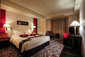 A hotel room with a large bed, two bedside tables with lamps, a desk with a chair, a TV, and a patterned carpet.