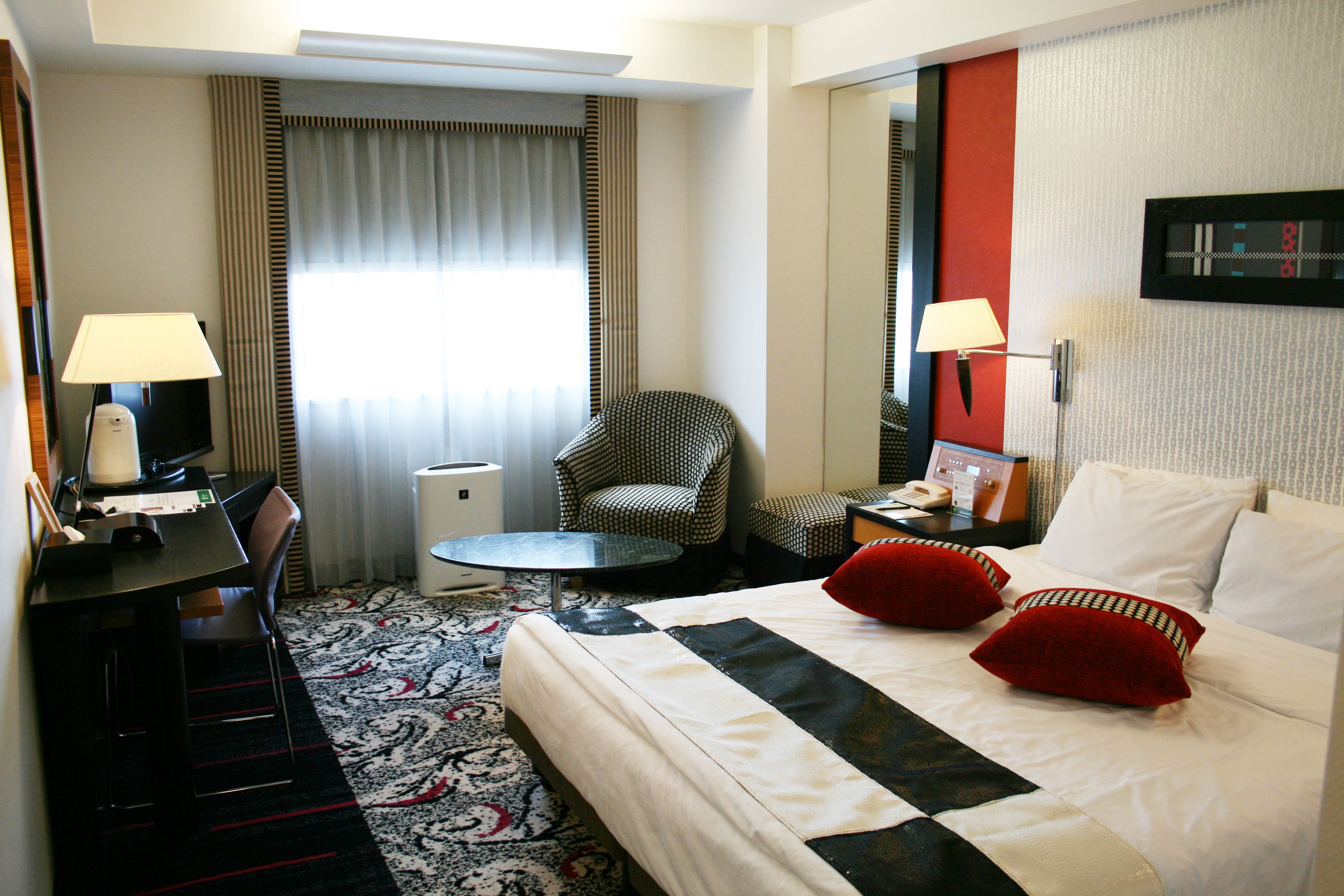 A hotel room with a bed, two red pillows, a desk with a lamp, a chair, a small table, and a mirror on the wall.