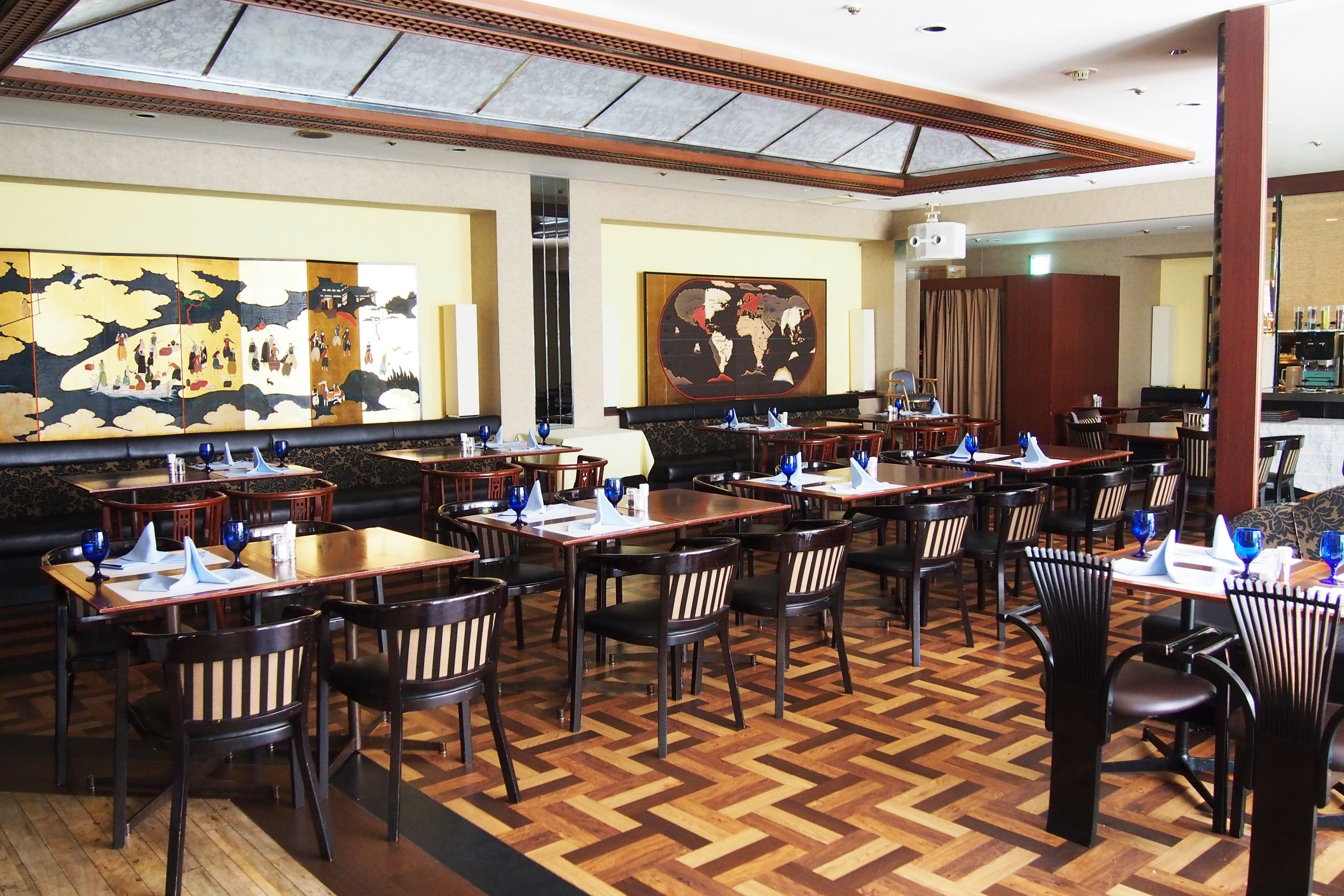 A restaurant with wooden tables and chairs, a herringbone floor pattern, and large wall art depicting traditional scenes.