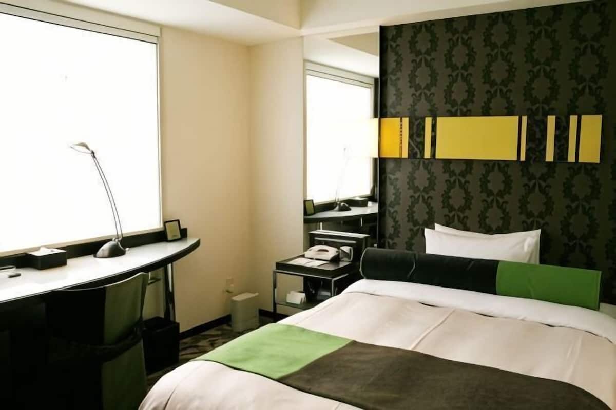 A hotel room with a bed, desk, chair, and a small table with a lamp.