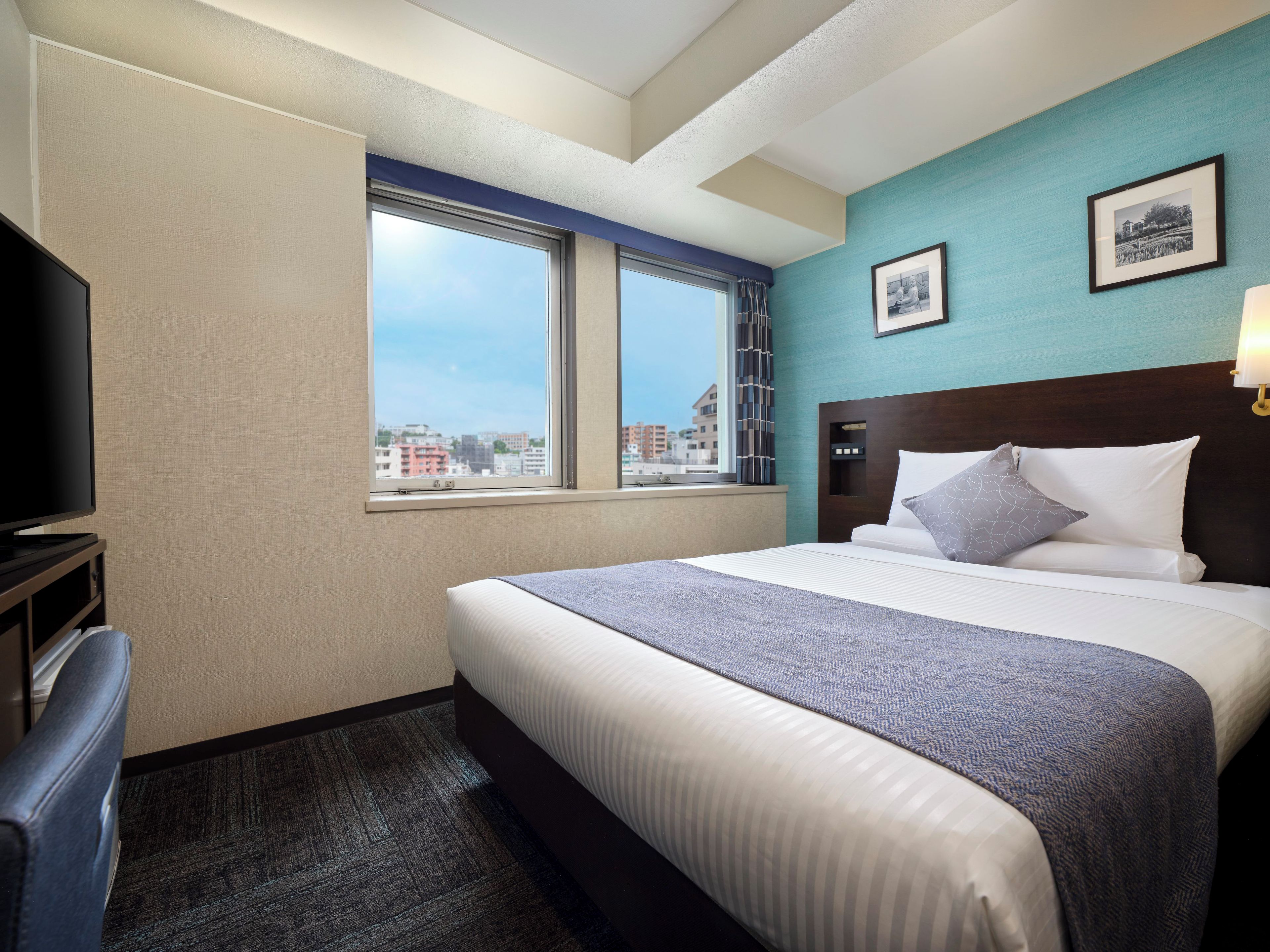A hotel room with a bed, a television, a window with a view of buildings, and a picture on the wall.