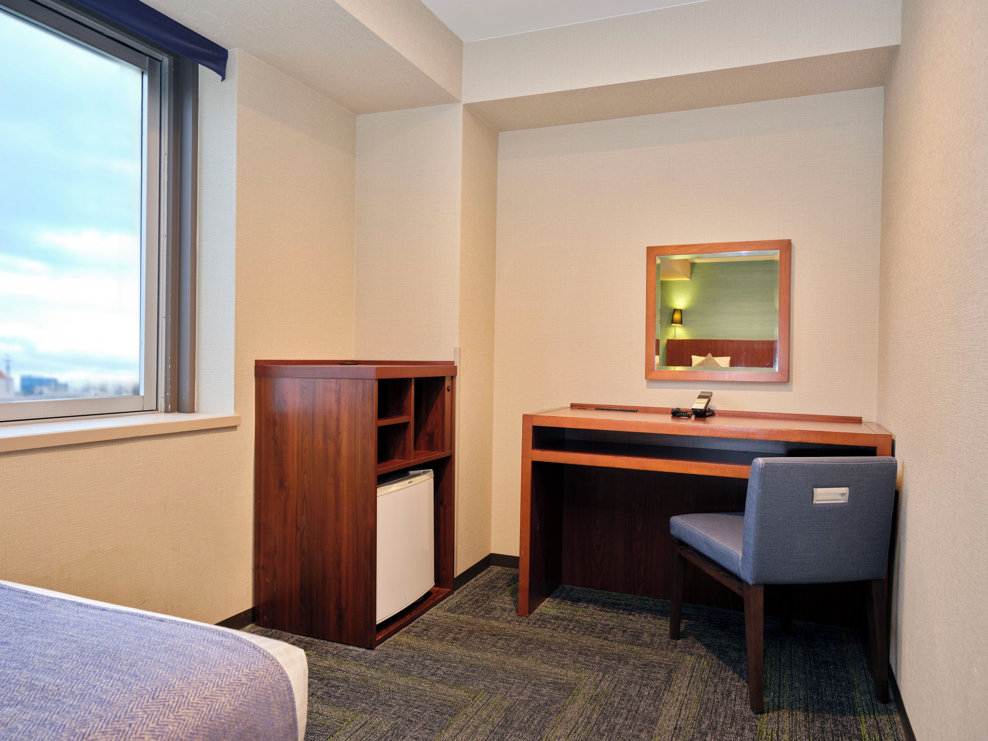 A hotel room with a bed, a desk, a chair, a small cabinet, and a window.