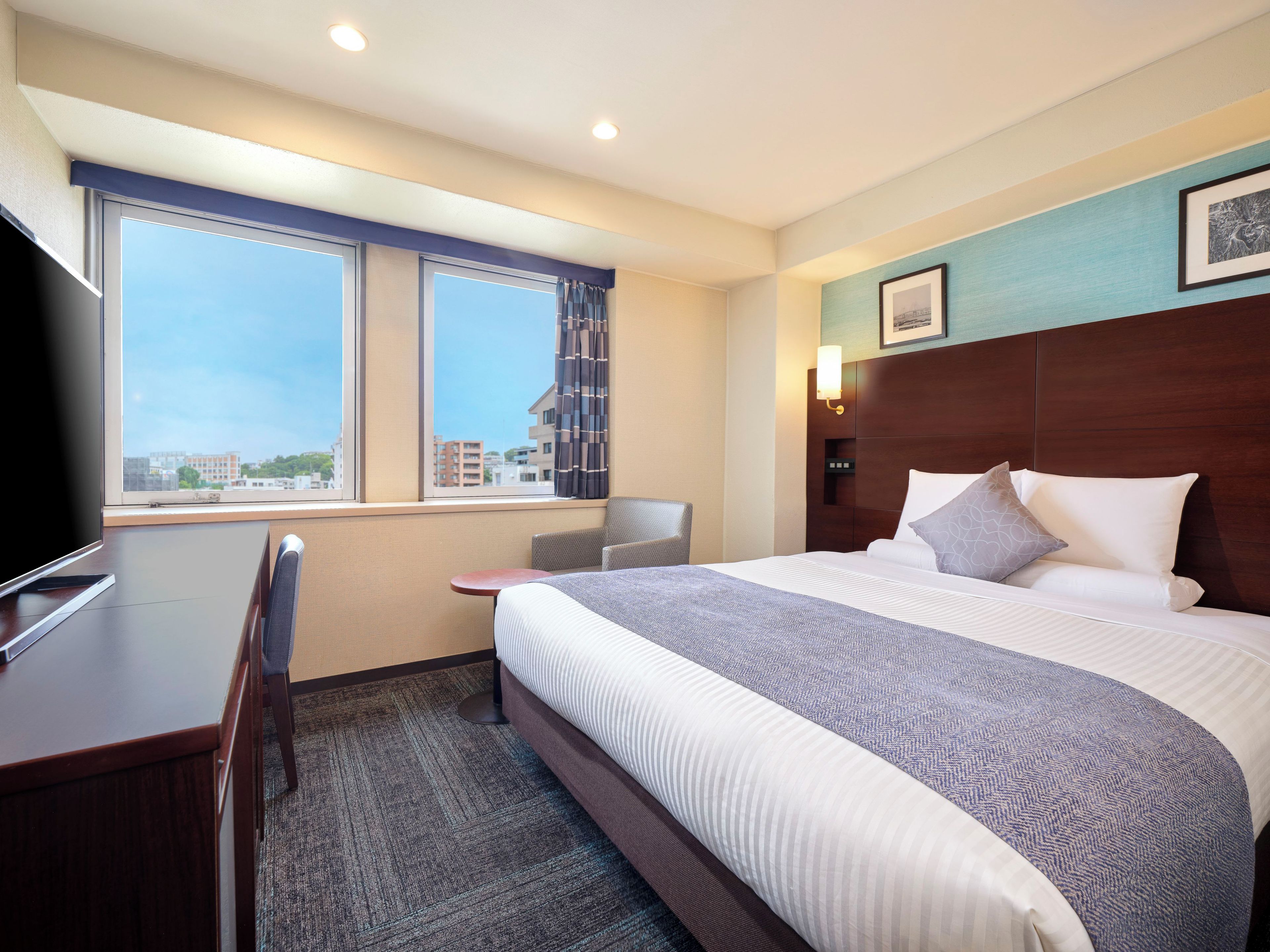 Ein Hotelzimmer mit Bett, Schreibtisch, Sessel, Fernseher, Fenster mit Vorhängen und Wandleuchte.