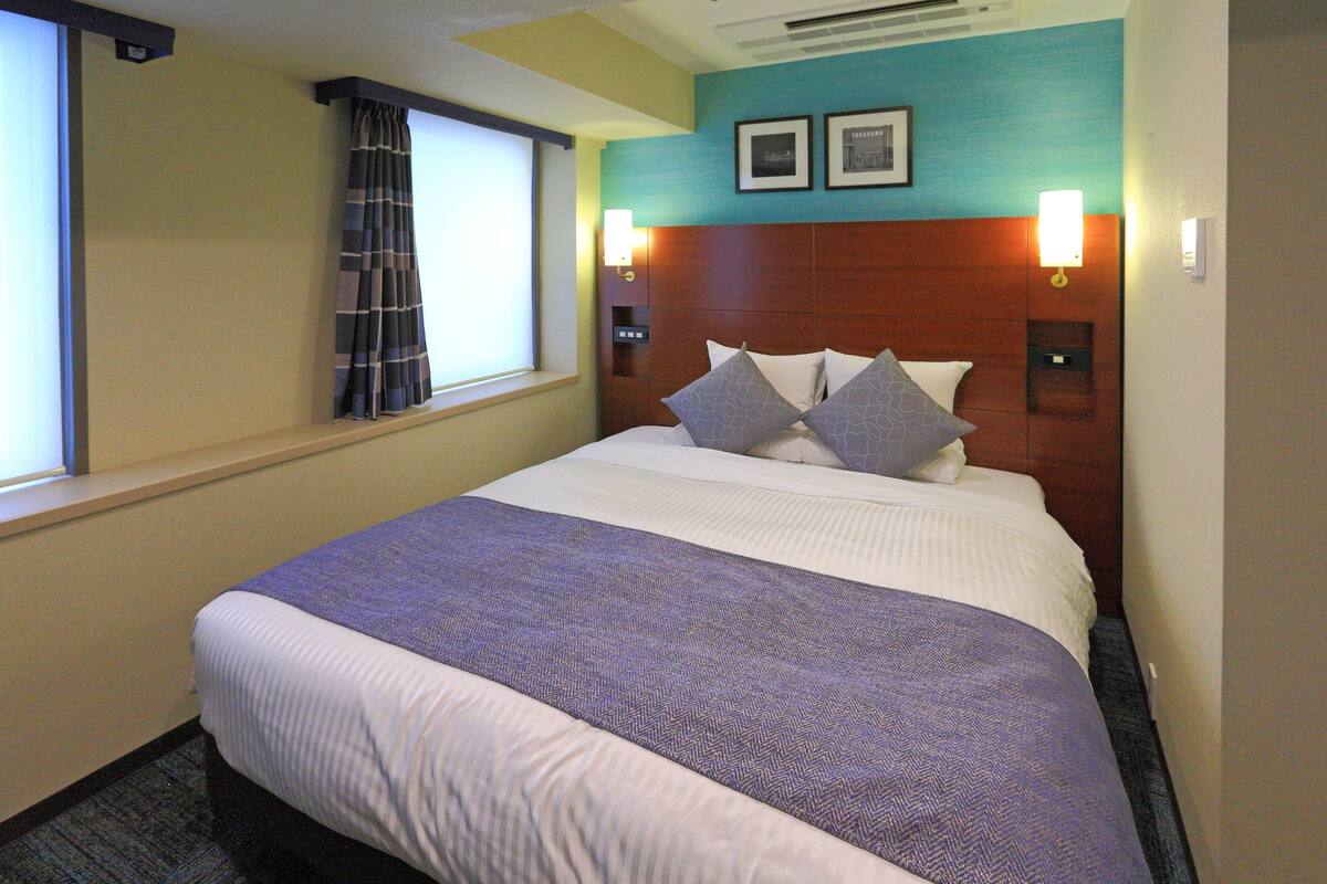 Een hotelkamer met een groot bed, een hoofdeinde met twee wandlampen, een raam met gordijn en een muur met twee ingelijste foto's.