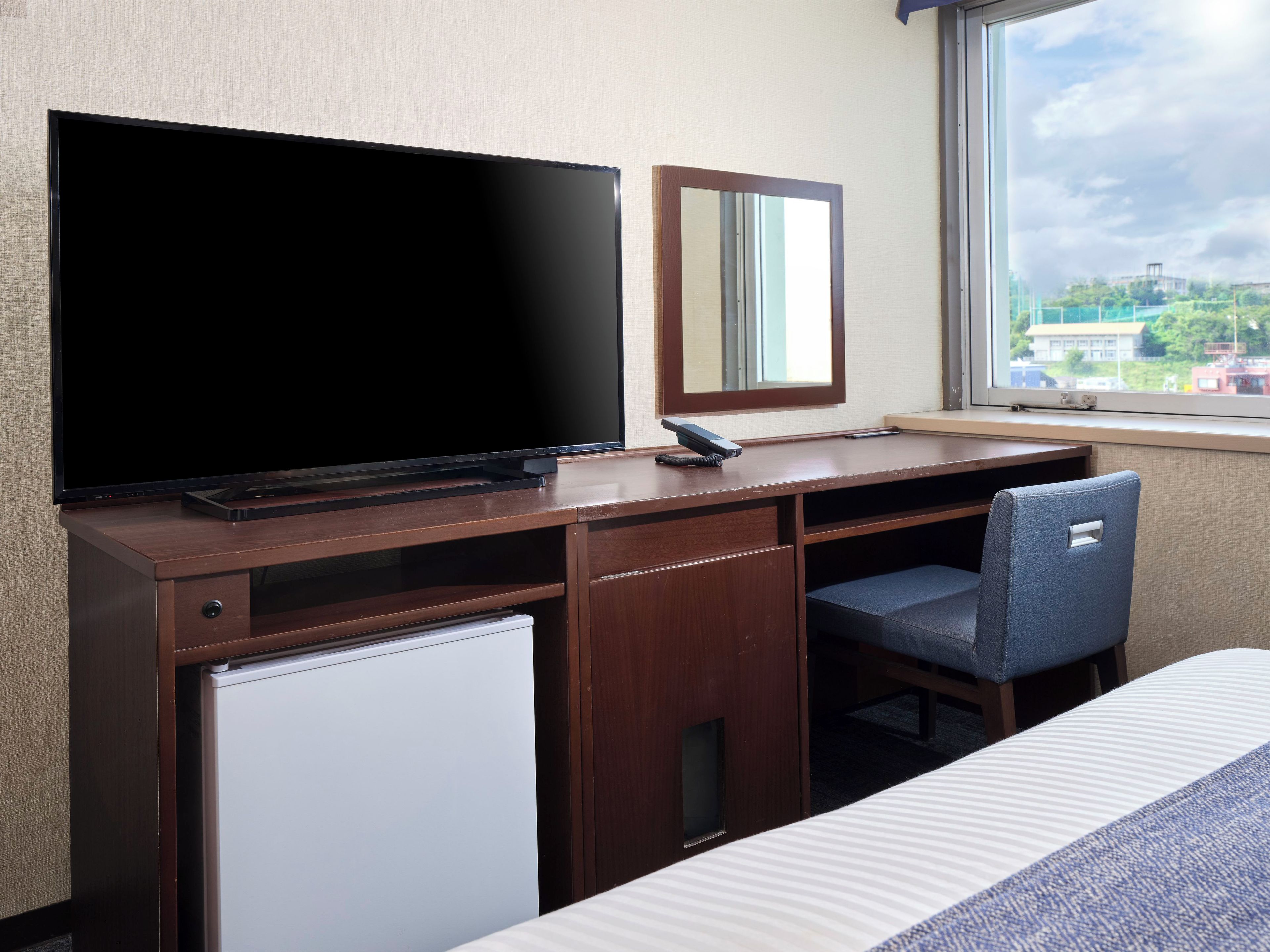 A hotel room with a bed, a desk with a computer, a TV, a mirror, and a window with a view of buildings.