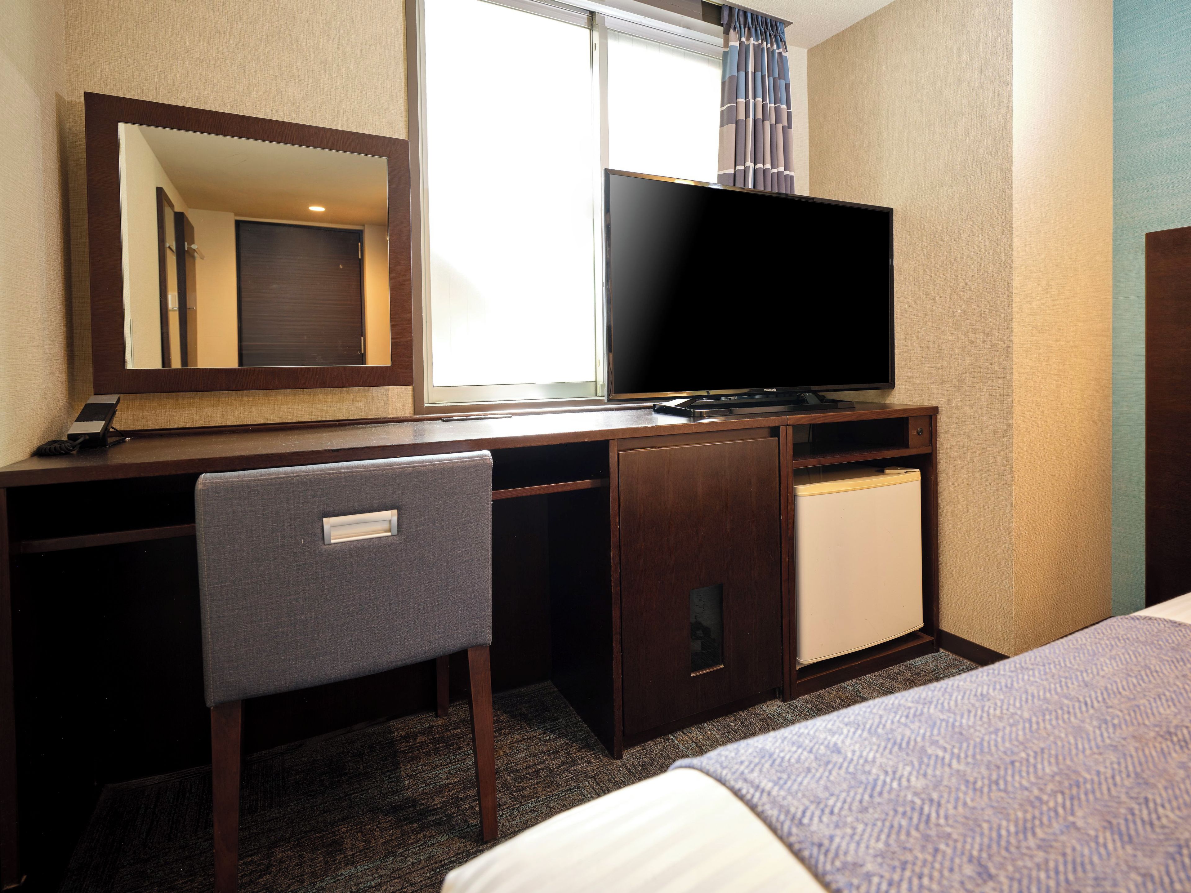 A hotel room with a bed, a desk with a television, a chair, and a small refrigerator.