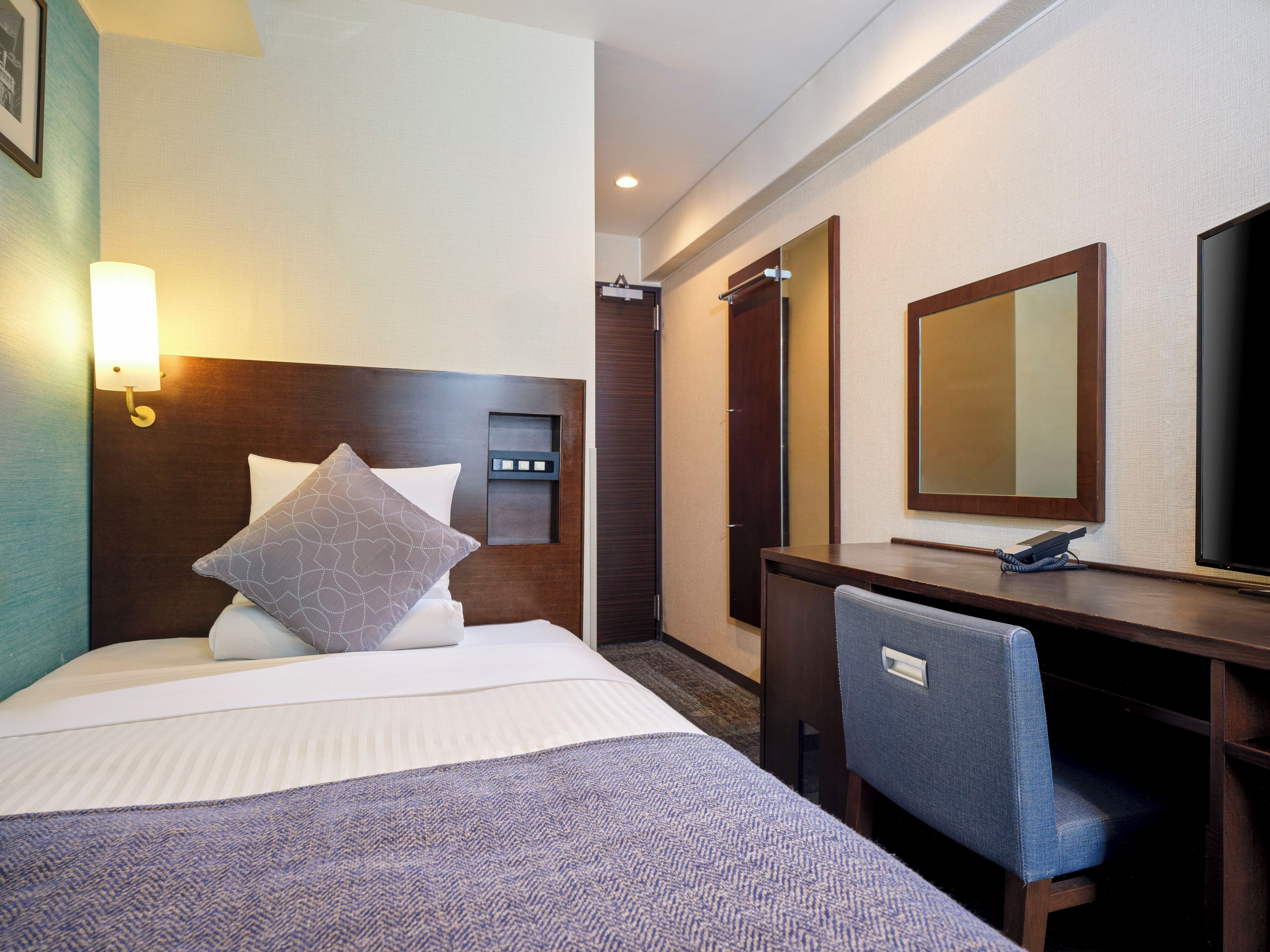 A hotel room with a bed, bedside table, desk, chair, and a large mirror.