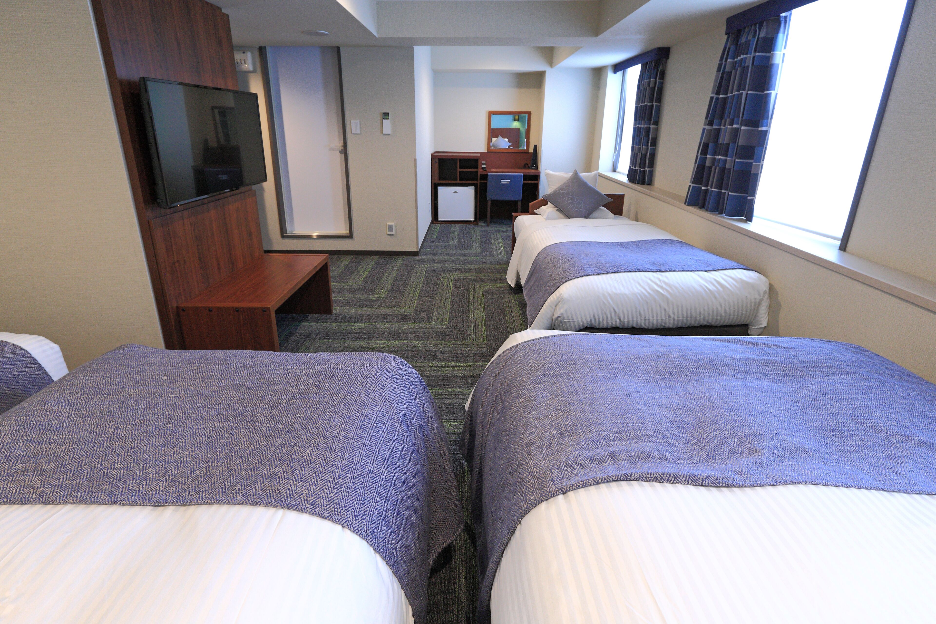 A hotel room with three beds, a TV, a desk, and a small refrigerator.