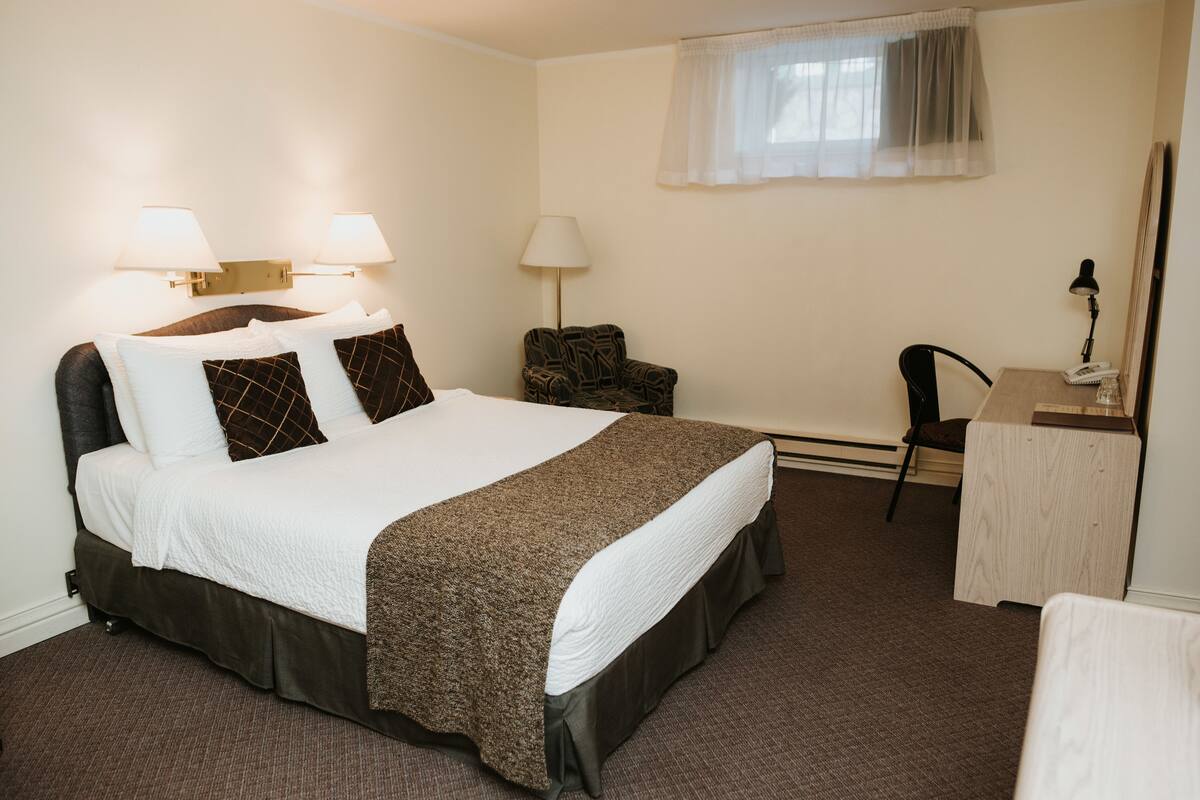 A hotel room with a large bed, two bedside lamps, a desk with a chair, and a window with a sheer curtain.