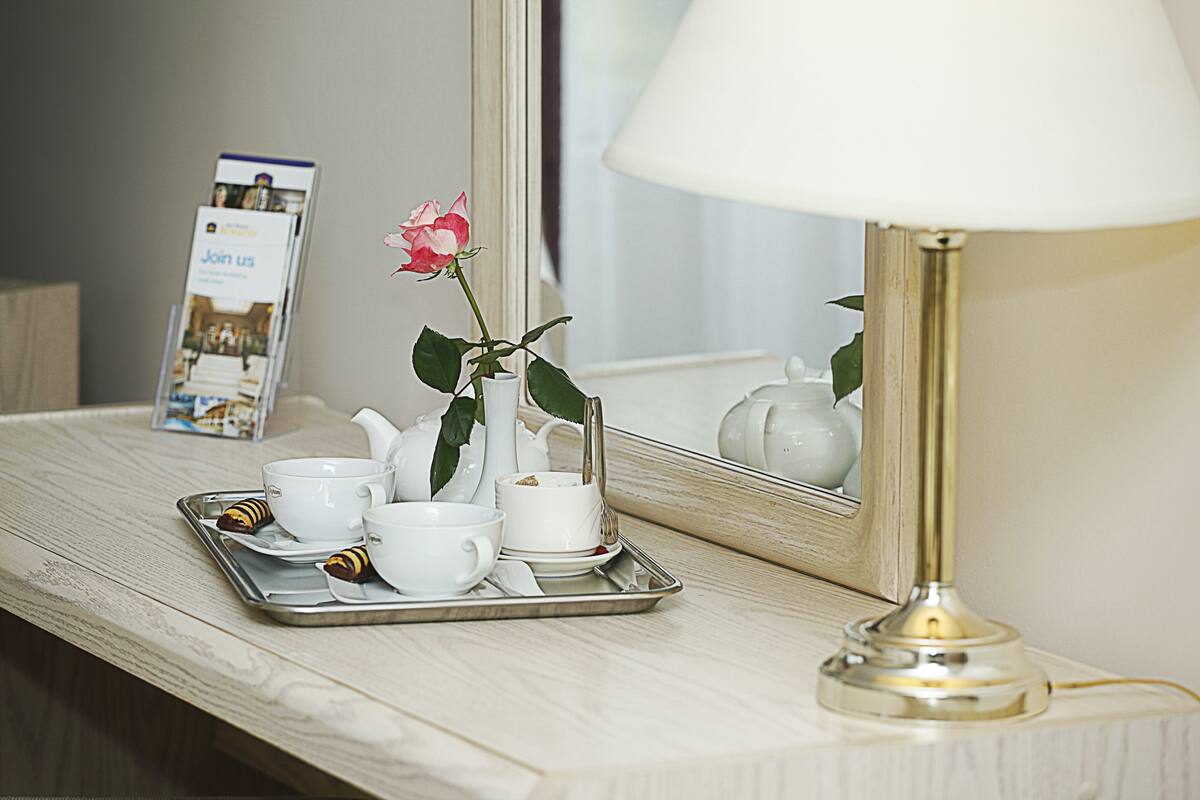 A tray with white cups and saucers, a teapot, and a rose on a wooden table with a lamp and a mirror reflecting the room.