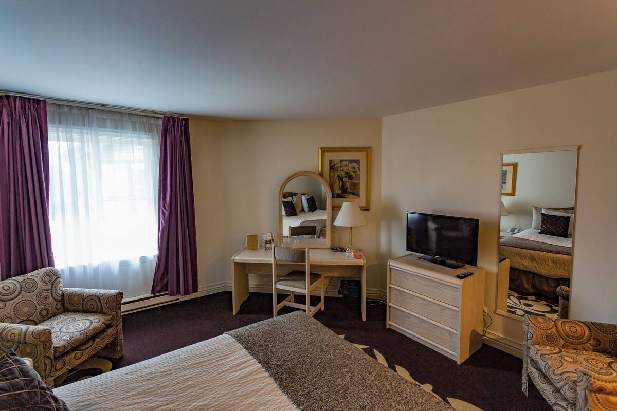 A hotel room with a bed, two chairs, a desk with a TV, a dresser, and a window with curtains.