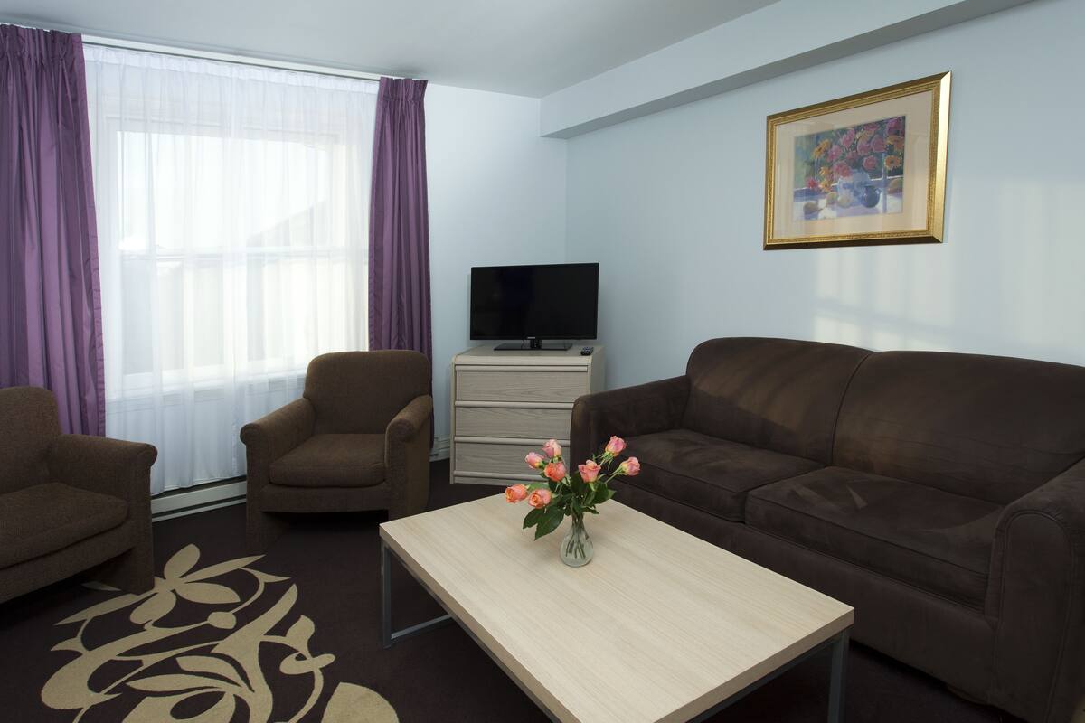 A living room with a sofa, two armchairs, a coffee table with a vase of flowers, a television on a stand, and a framed picture on the wall.