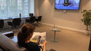 A hotel lobby with a large TV screen displaying a cityscape, a seating area with a sofa and chairs, and a small table with a plant. - City Hotel (Luxembourg City)
