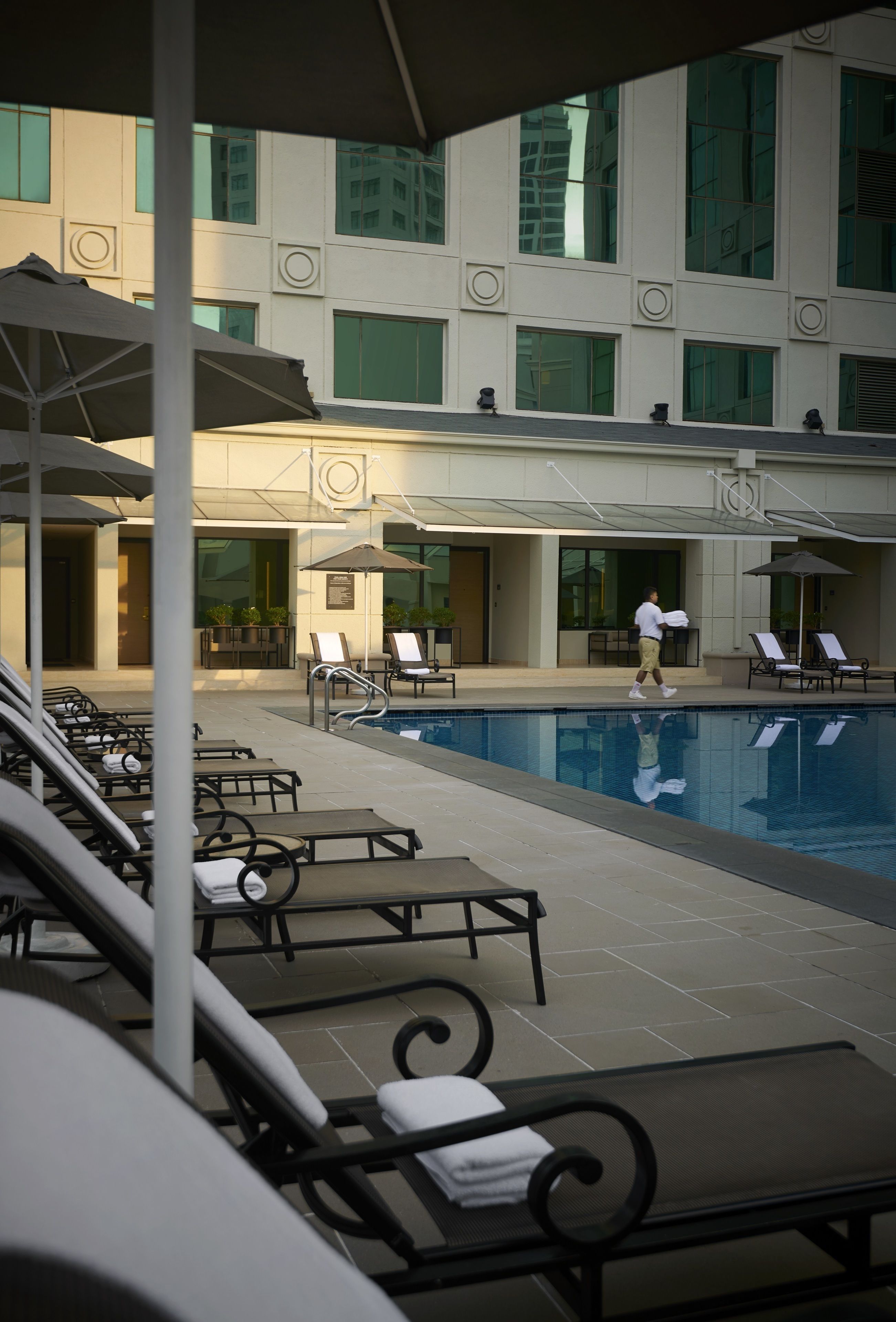 A poolside area with lounge chairs and umbrellas, a pool, and a building with large windows and a flat roof.