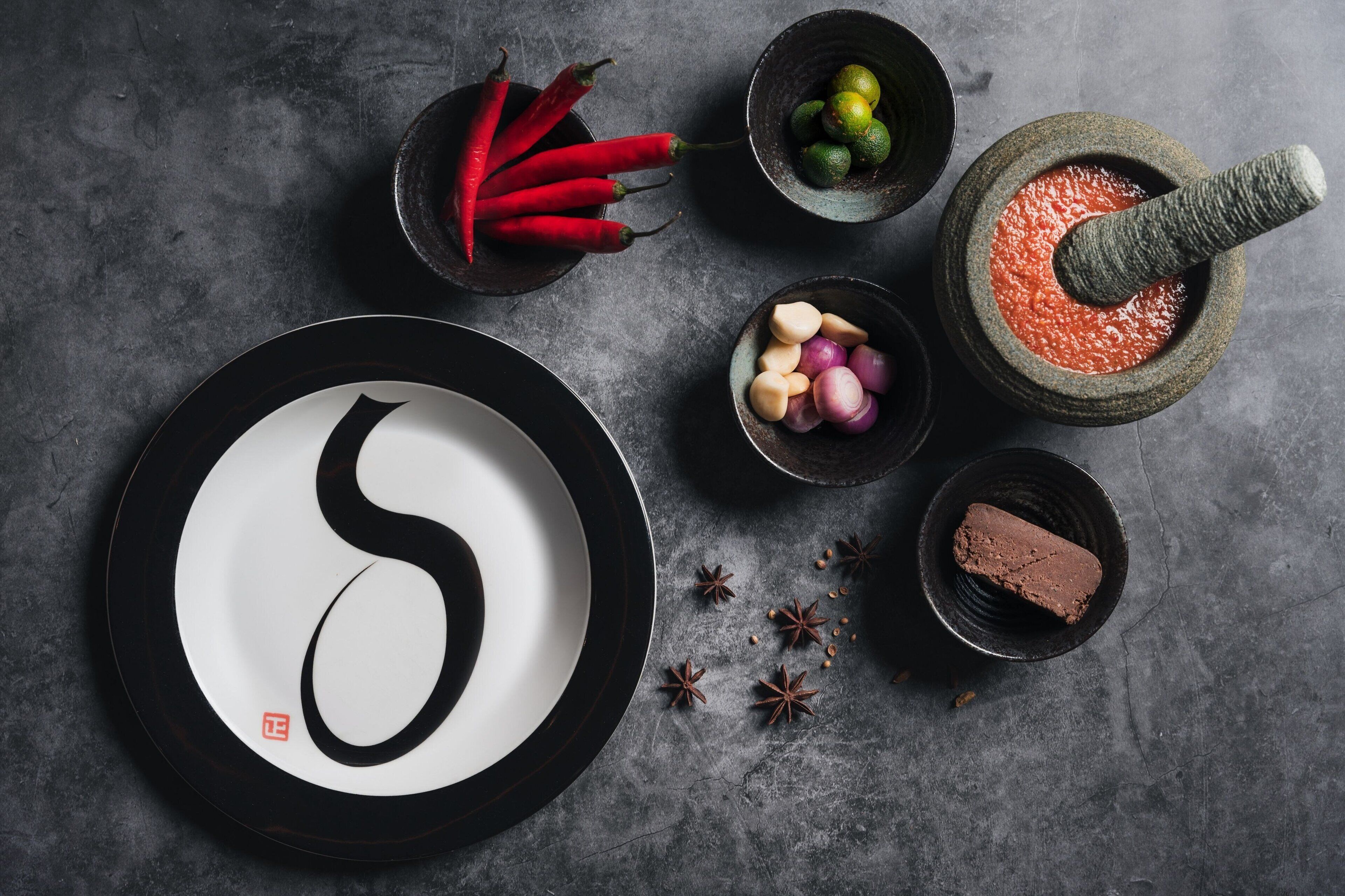 A black and white plate with a red and white design, surrounded by various food items and spices on a textured surface.