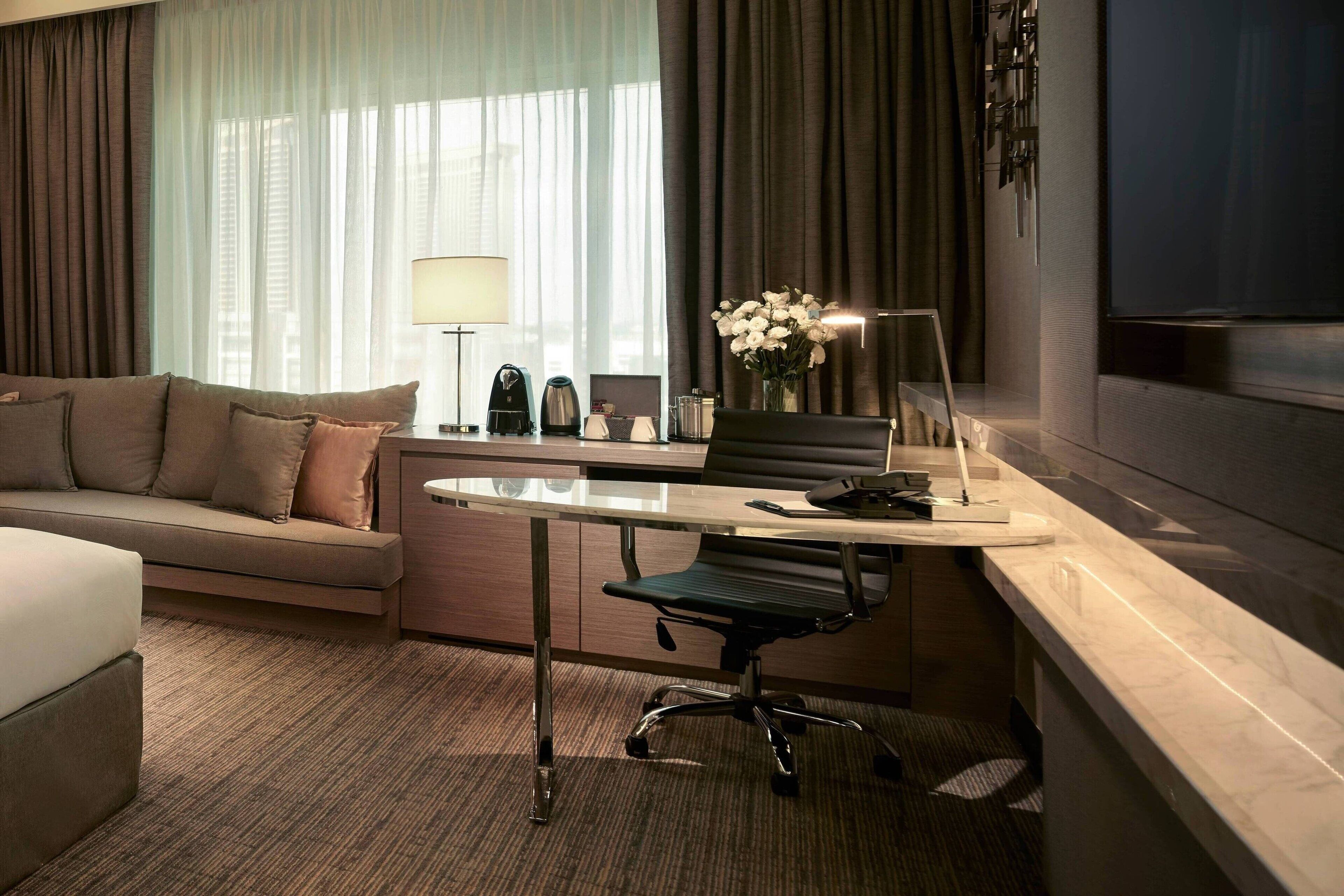 A hotel room with a sofa, a desk with a chair, a lamp, a vase with flowers, and a TV.