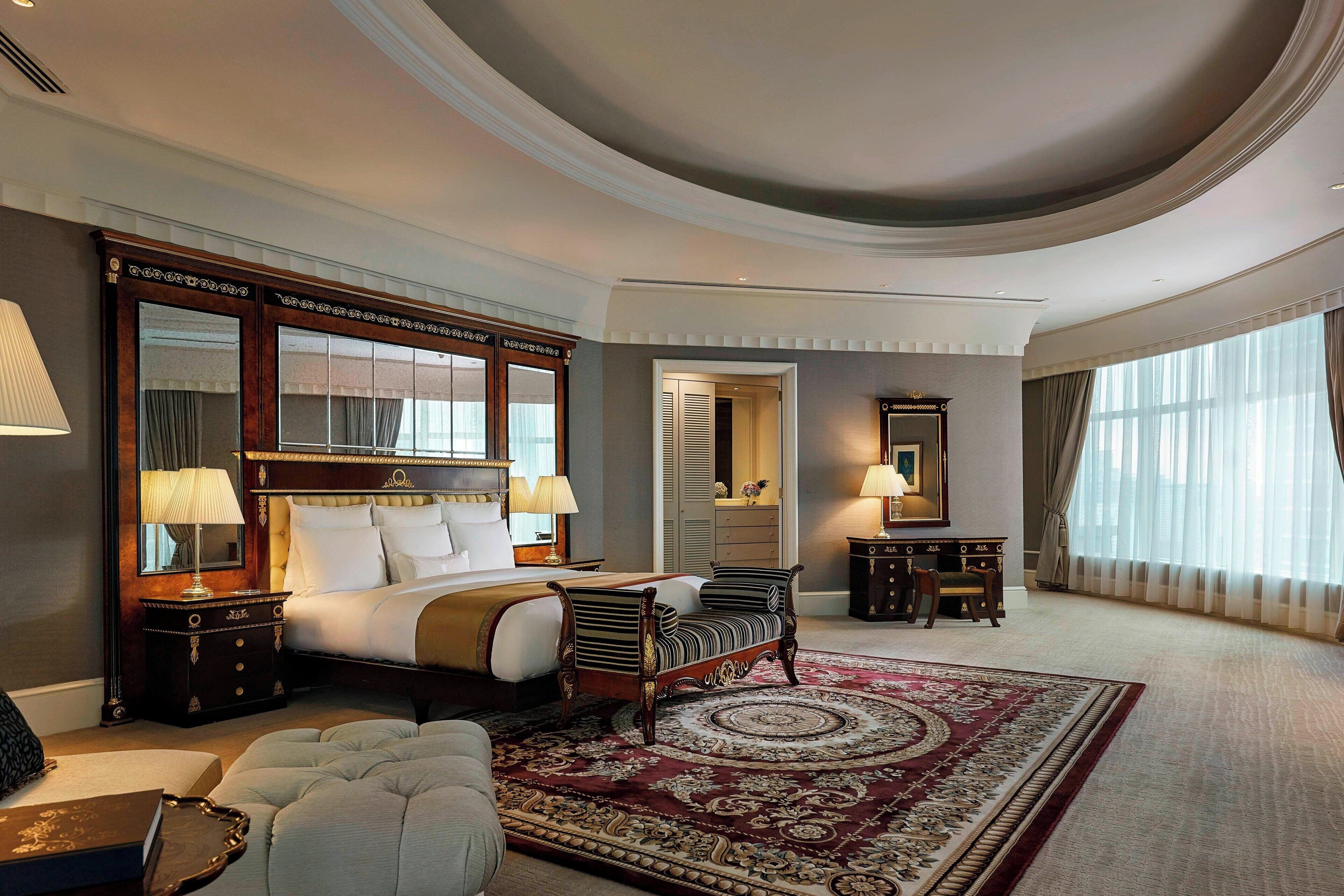 A spacious hotel room with a large bed, a sitting area, a decorative rug, and a view of the outdoors through large windows.