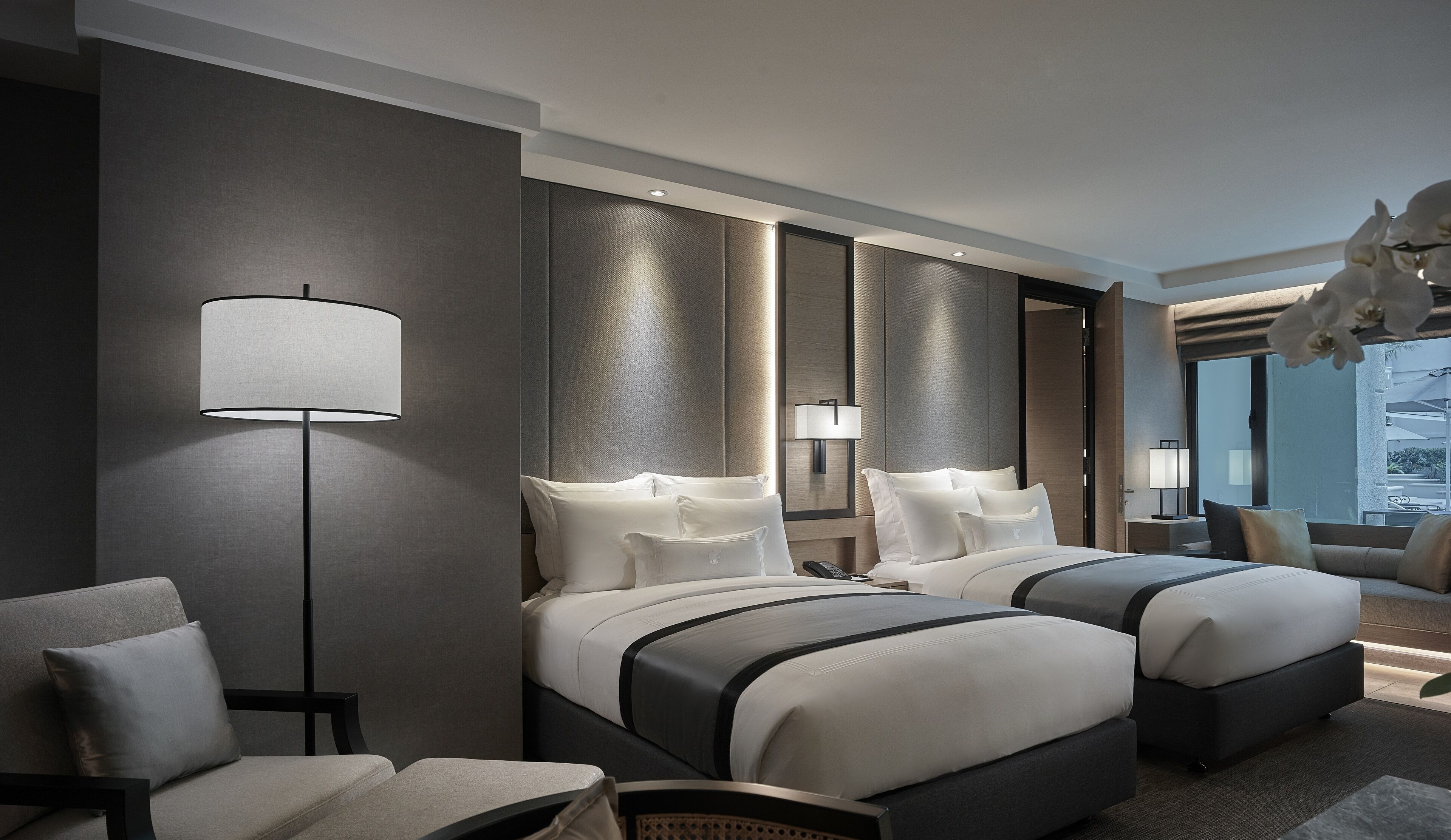 Una habitación de hotel moderna con dos camas, un rincón de estar con sofá y sillones, una lámpara de pie y un ventanal con vistas.