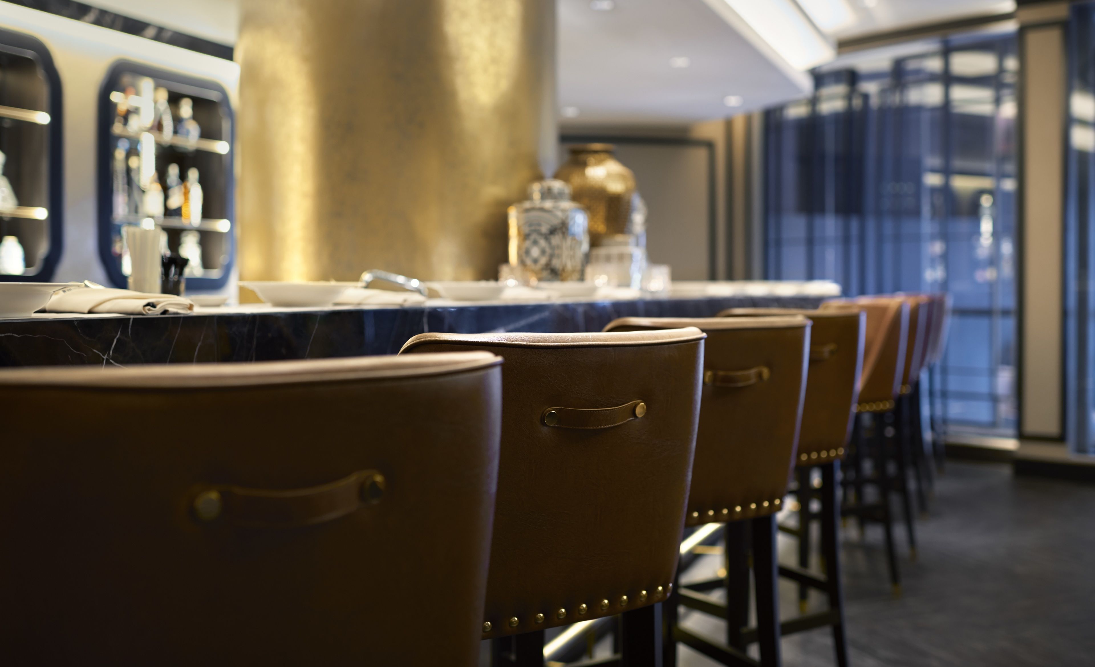 Una hilera de taburetes de cuero marrón con patas de metal, una mesa con cubierta de mármol y una zona de bar con botellas y un jarrón decorativo.