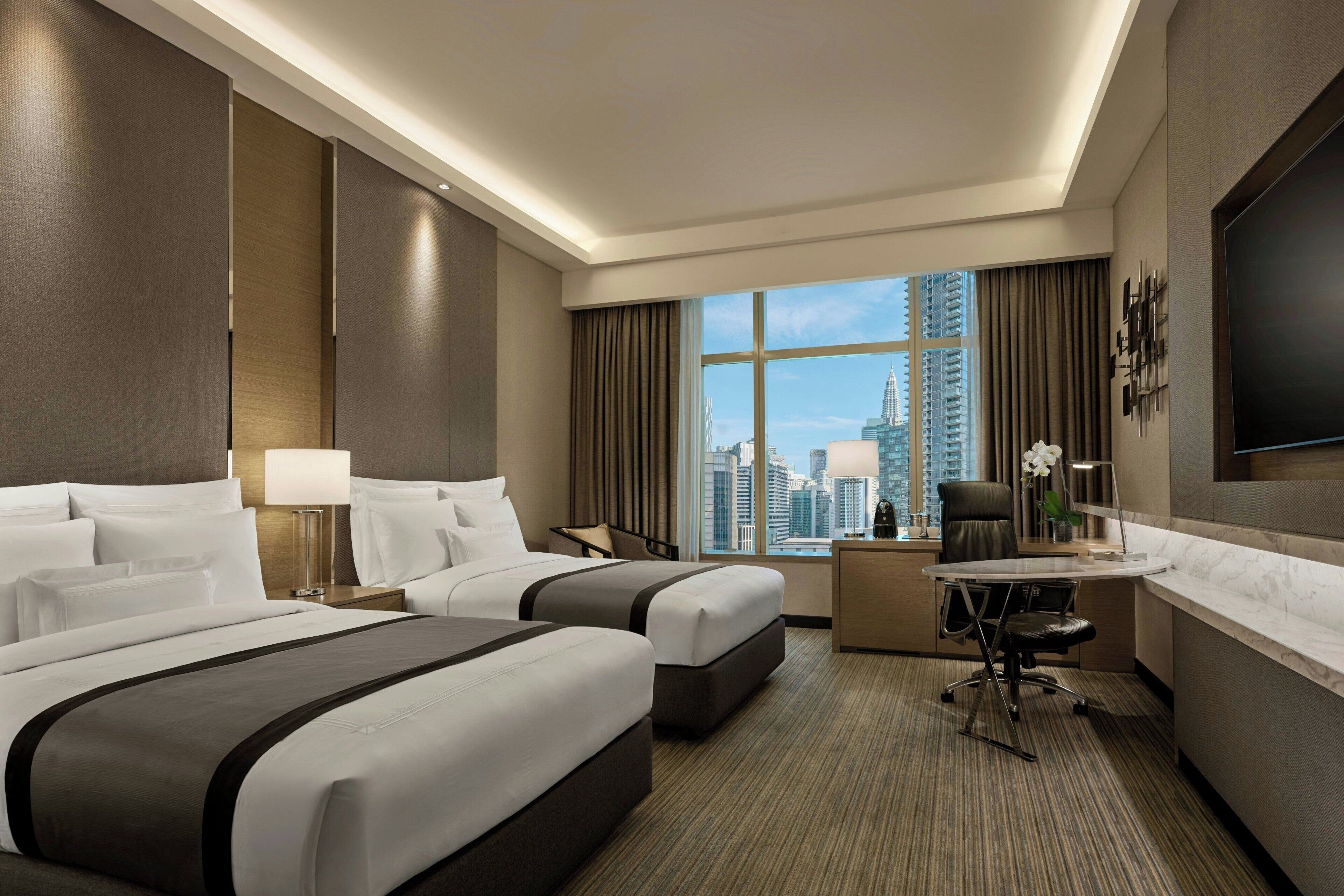 A hotel room with two beds, a desk, a chair, a TV, and a view of the cityscape.