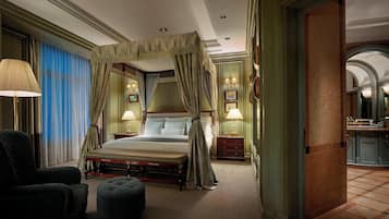 A bedroom with a four-poster bed, a sitting area with a chair and ottoman, and a bathroom visible through an open doorway.