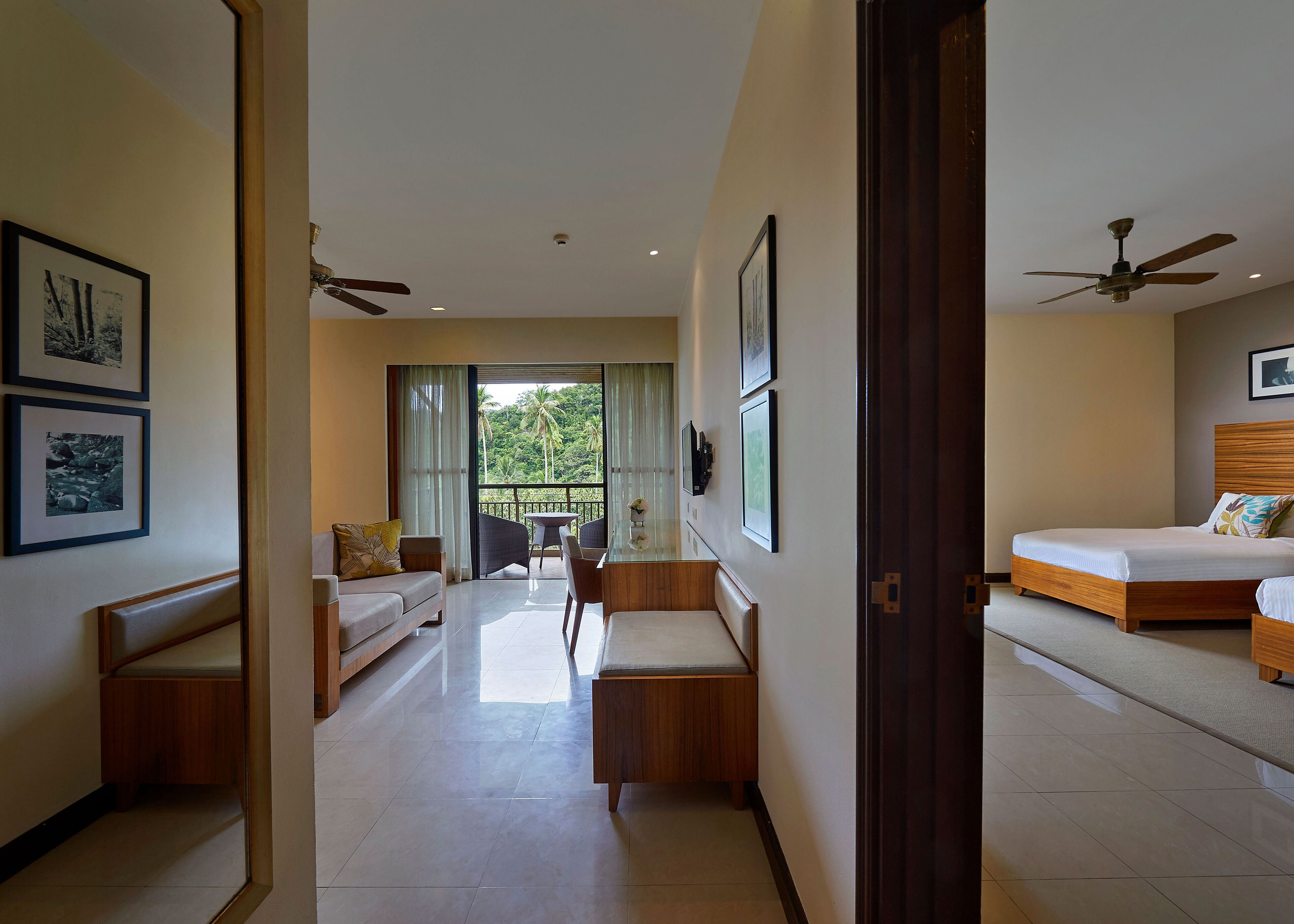 A hotel room with a bed, a sofa, a dining table, and a balcony with a view of greenery.