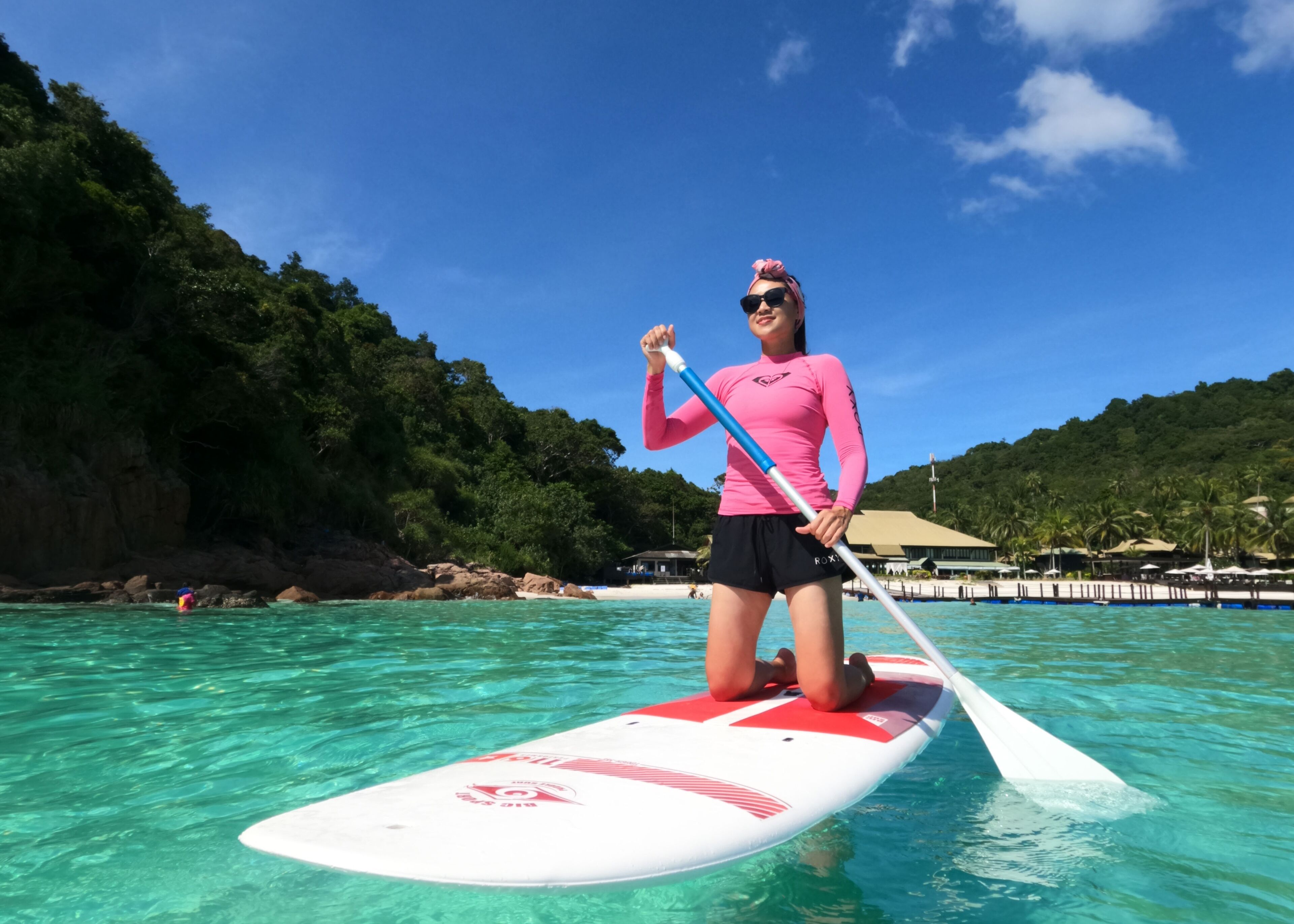 A person standing on a paddleboard in clear water with a tropical beach and lush greenery in the background.