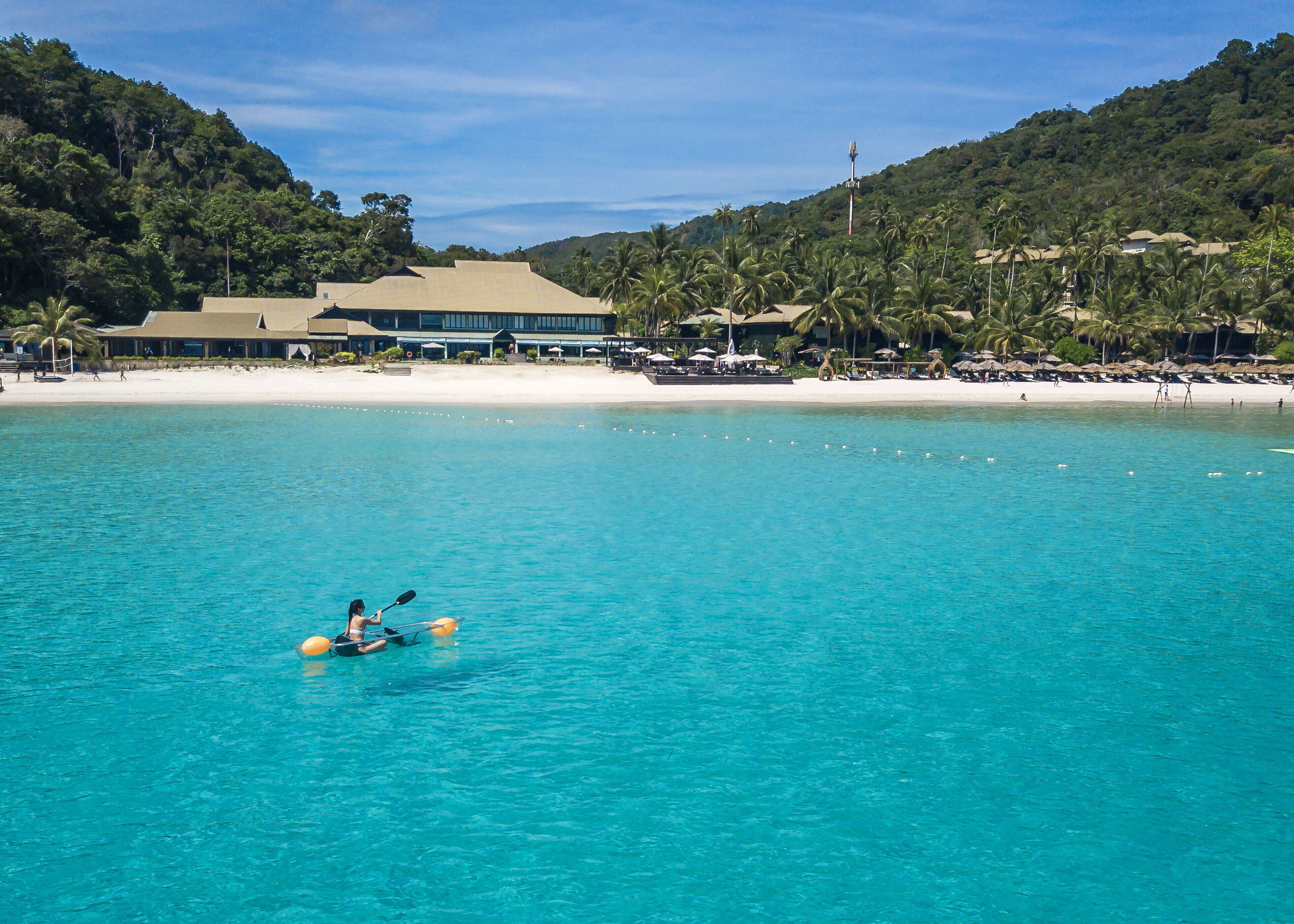 A coastal resort with a clear turquoise sea, a sandy beach, and a building with a flat roof.