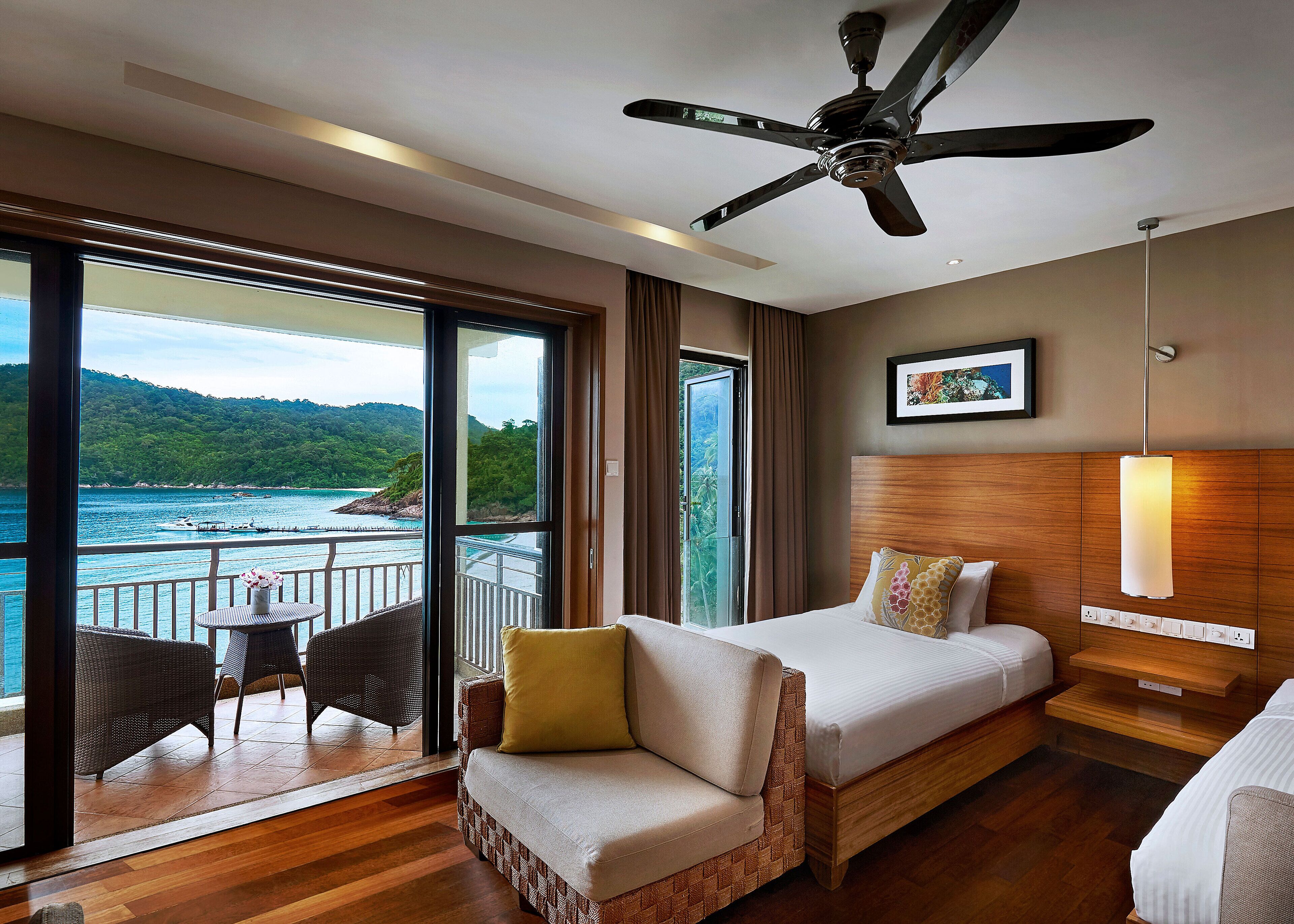 A hotel room with a bed, a sofa, a ceiling fan, a large window with a view of the sea, and a small table with chairs.