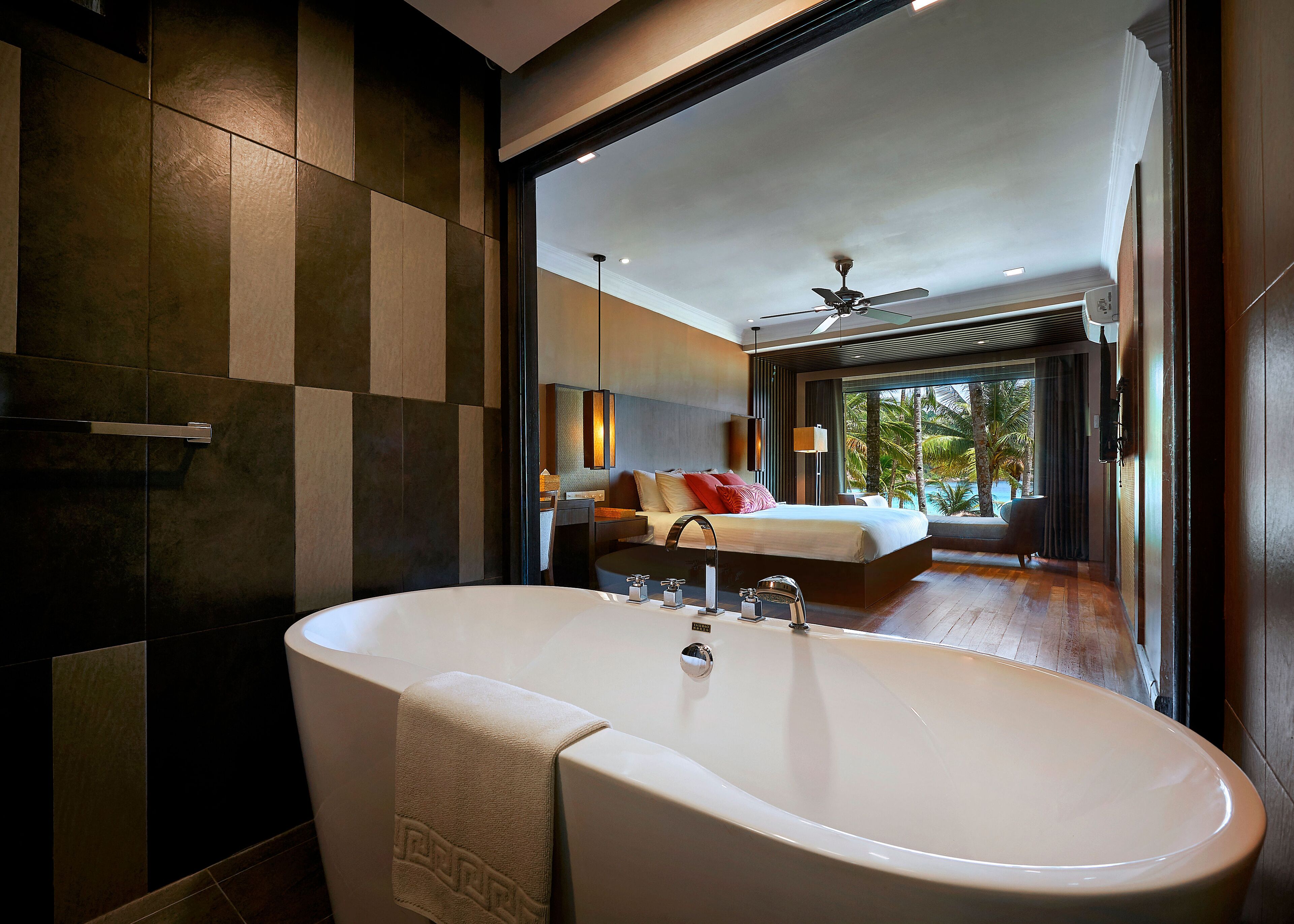 A modern bathroom with a large bathtub, a visible towel, and a view of a bedroom with a bed and a window.
